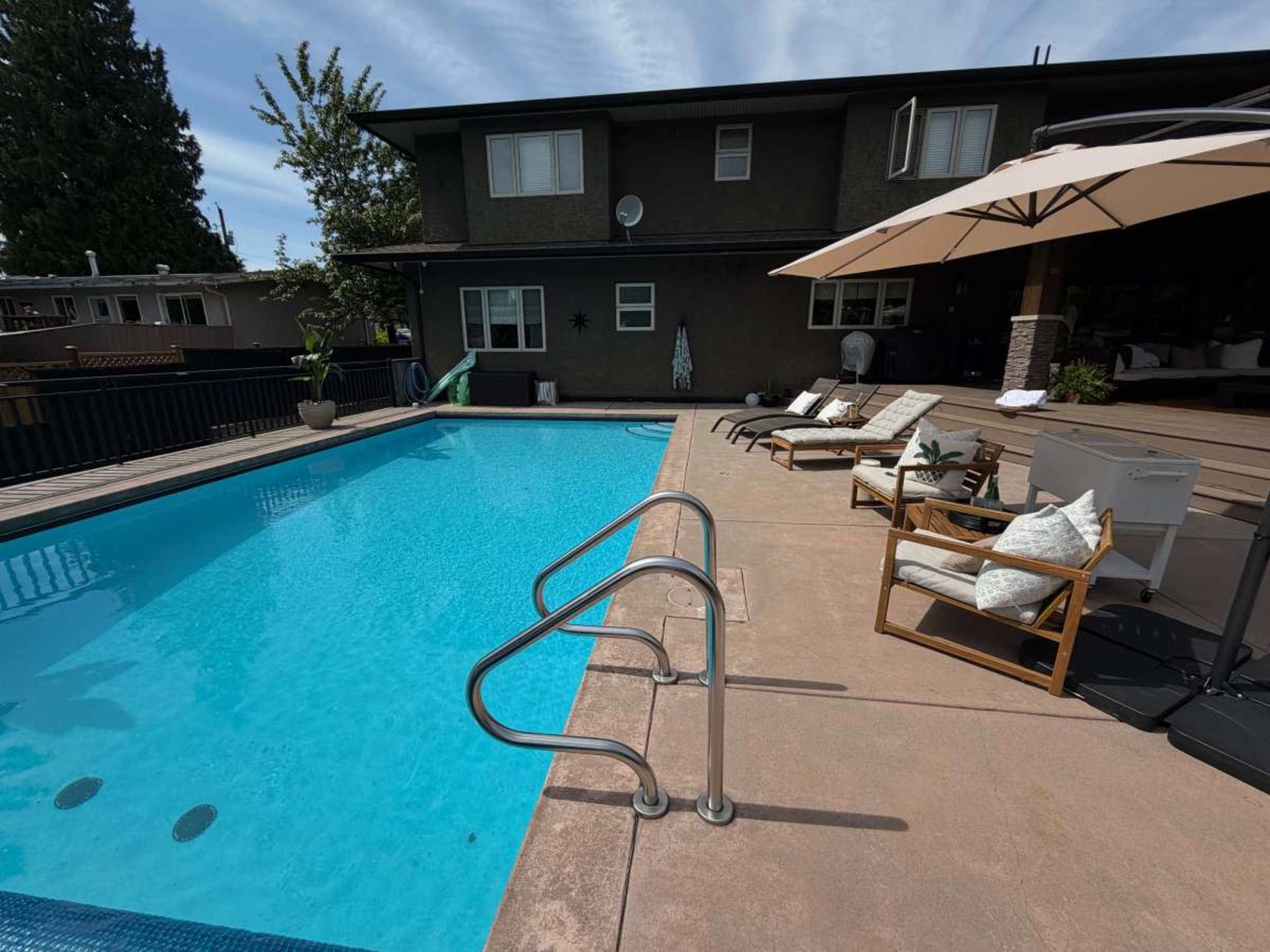 A clear swimming pool with steps leads to a deck area featuring lounge chairs and an umbrella near a building with multiple windows.