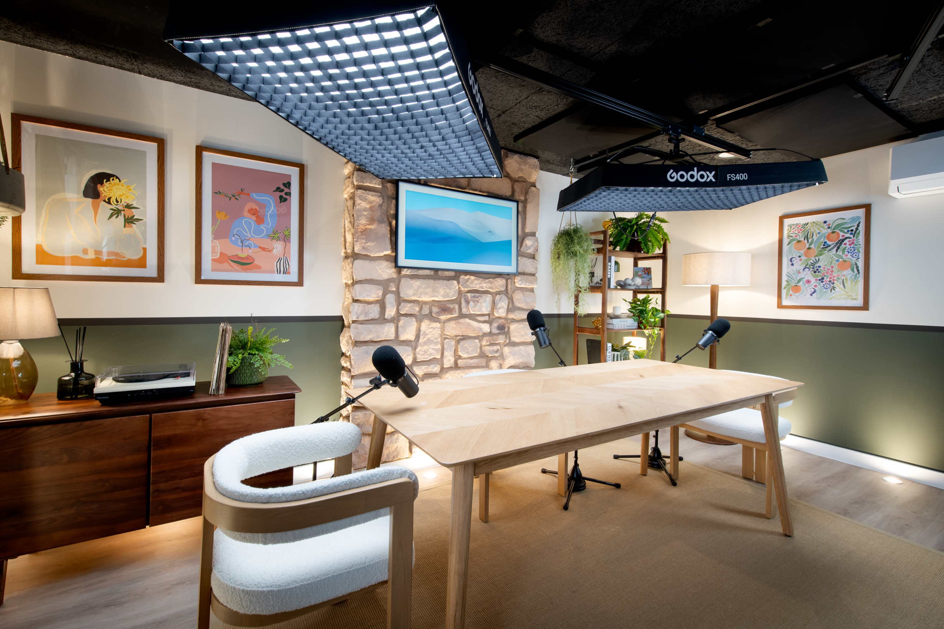 The image shows a modern podcast studio with a wooden table, two chairs, and microphones, accentuated by framed artwork and a stone wall.