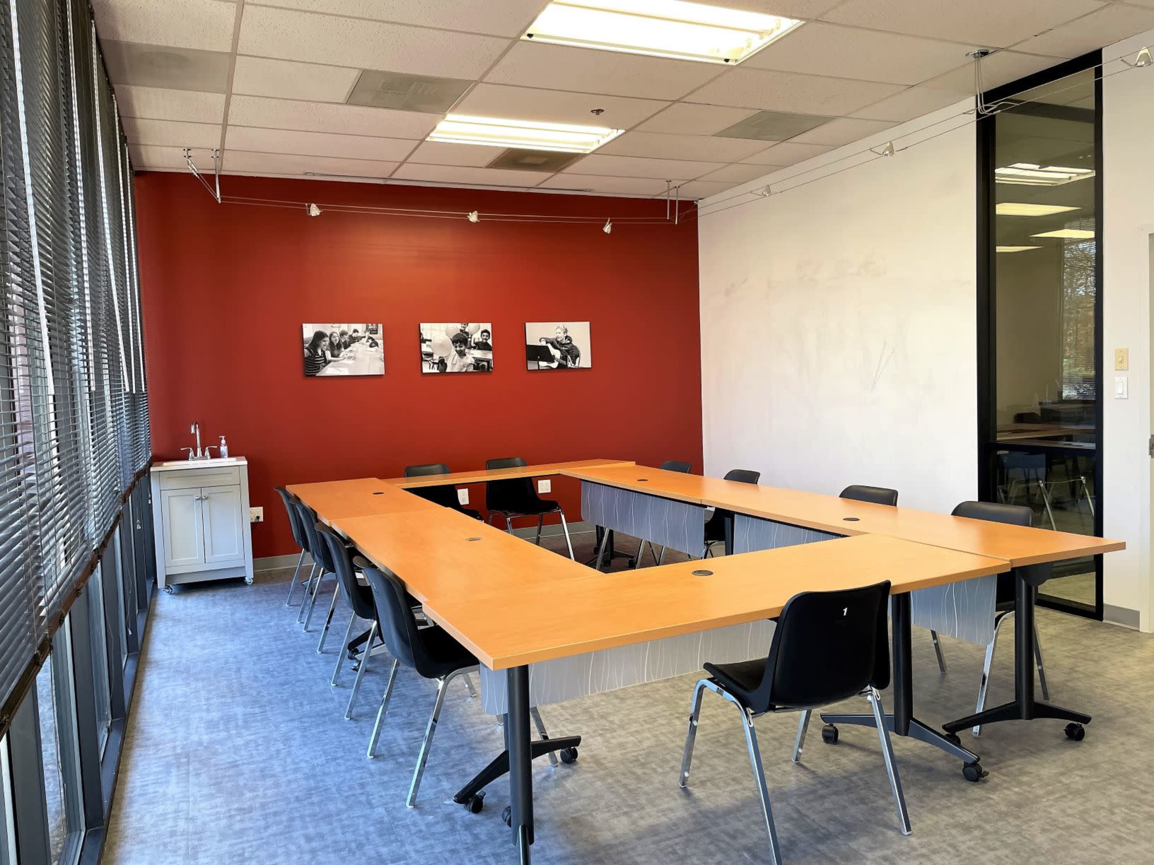 A conference room featuring a rectangular table surrounded by black chairs, with a red accent wall displaying three black-and-white photographs.