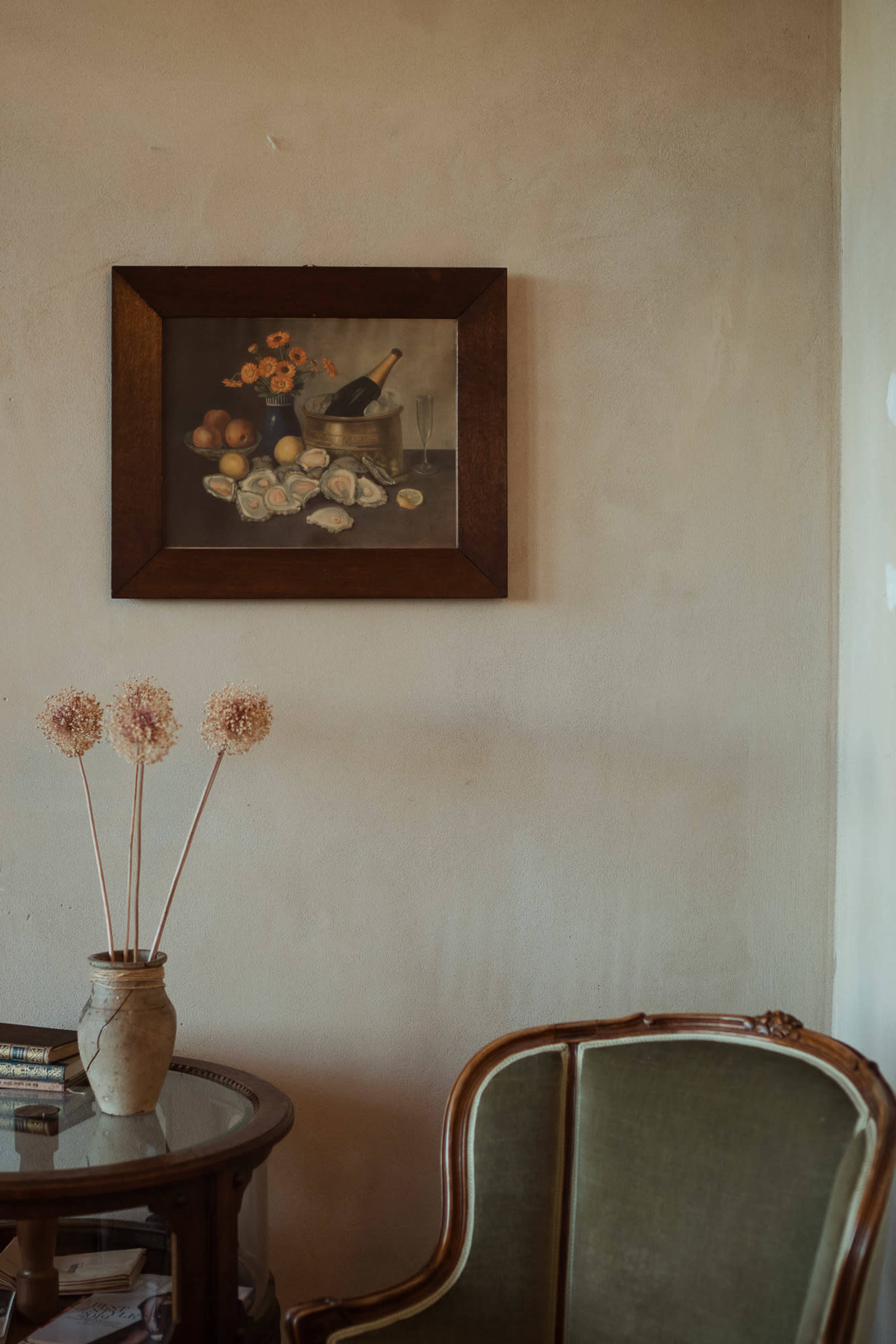 A still life painting of flowers and fruits hangs on a plaster wall above a small glass table and an upholstered chair.
