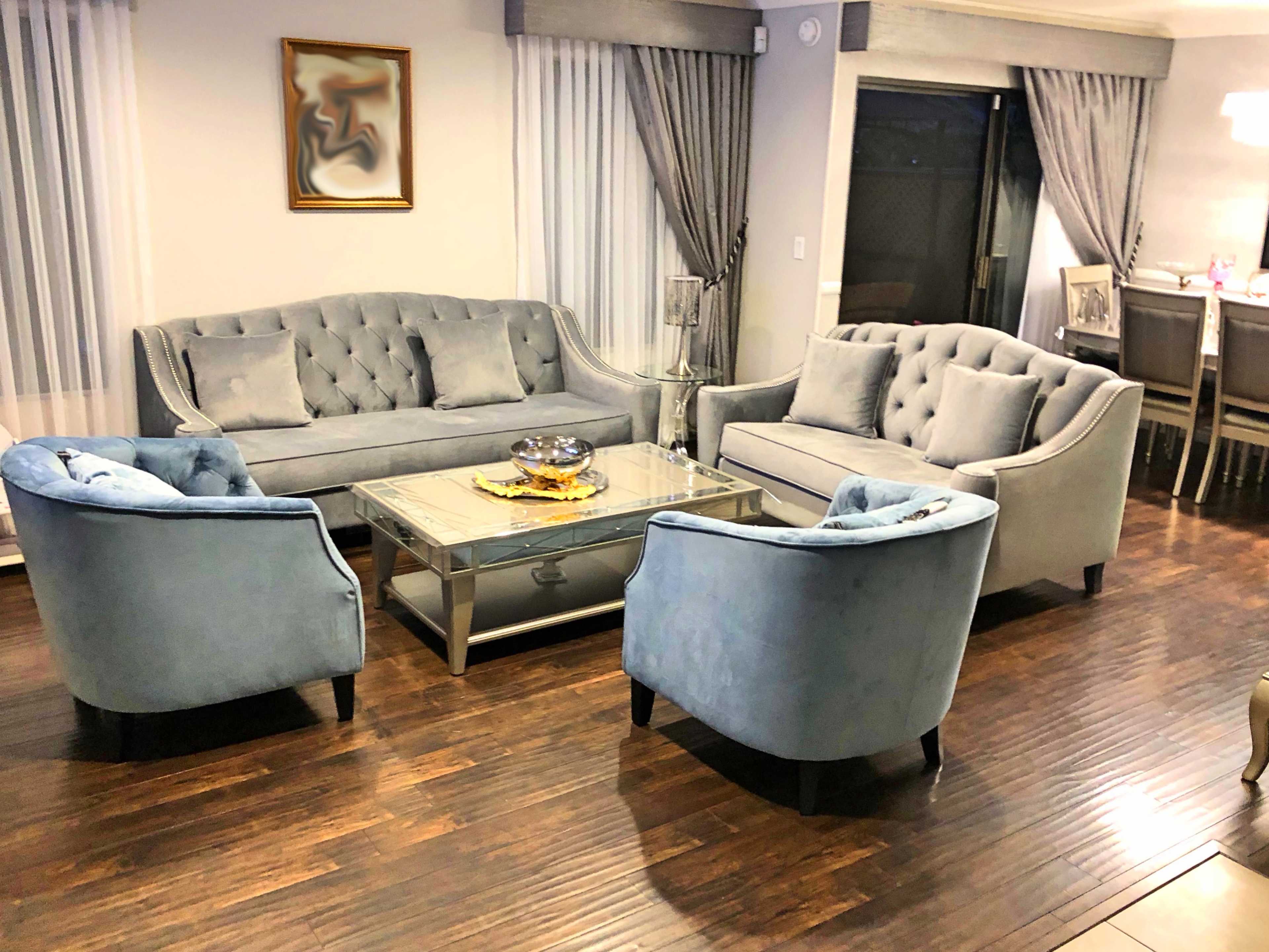 A living room with a gray tufted sofa and two matching chairs arranged around a glass coffee table on a hardwood floor, with a dining area visible in the background.