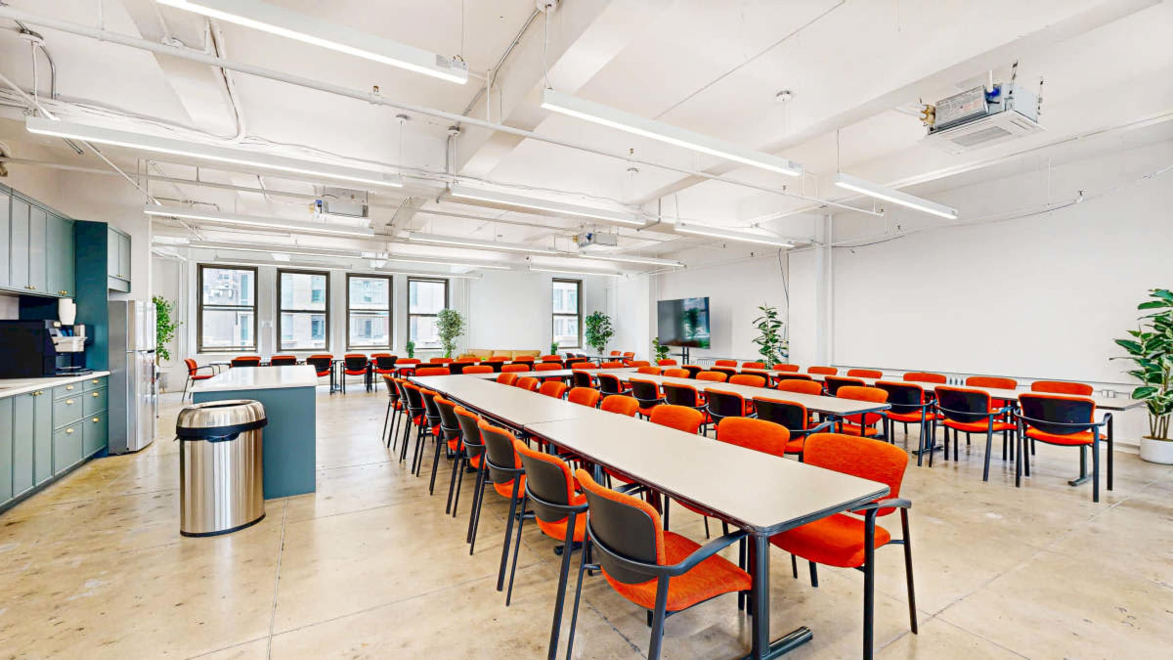 A spacious meeting room features long tables arranged in rows, surrounded by red chairs, with windows and greenery visible in the background.
