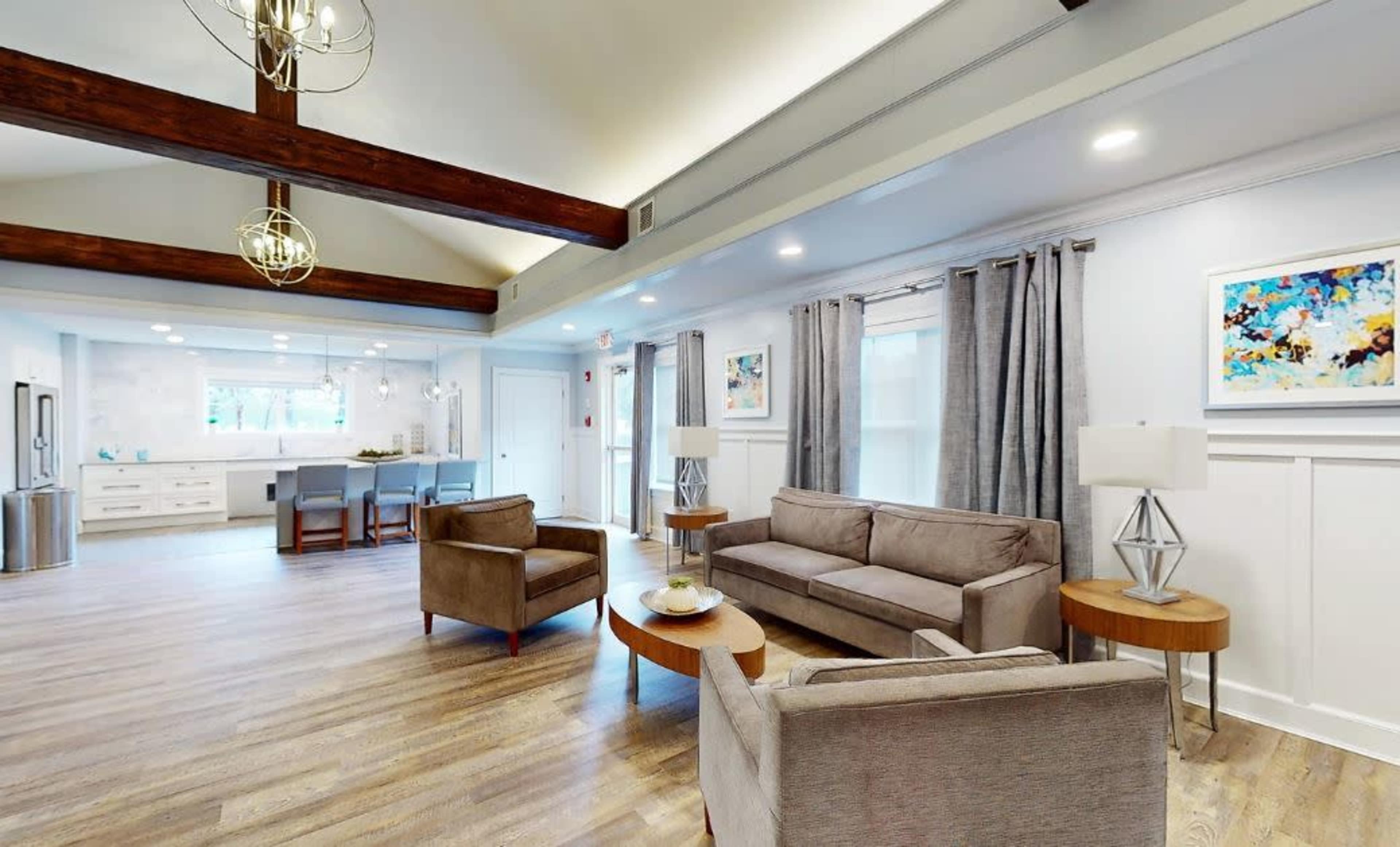 The image shows a modern, well-lit living space with gray furniture, wooden beams on the ceiling, and a kitchen area visible in the background.