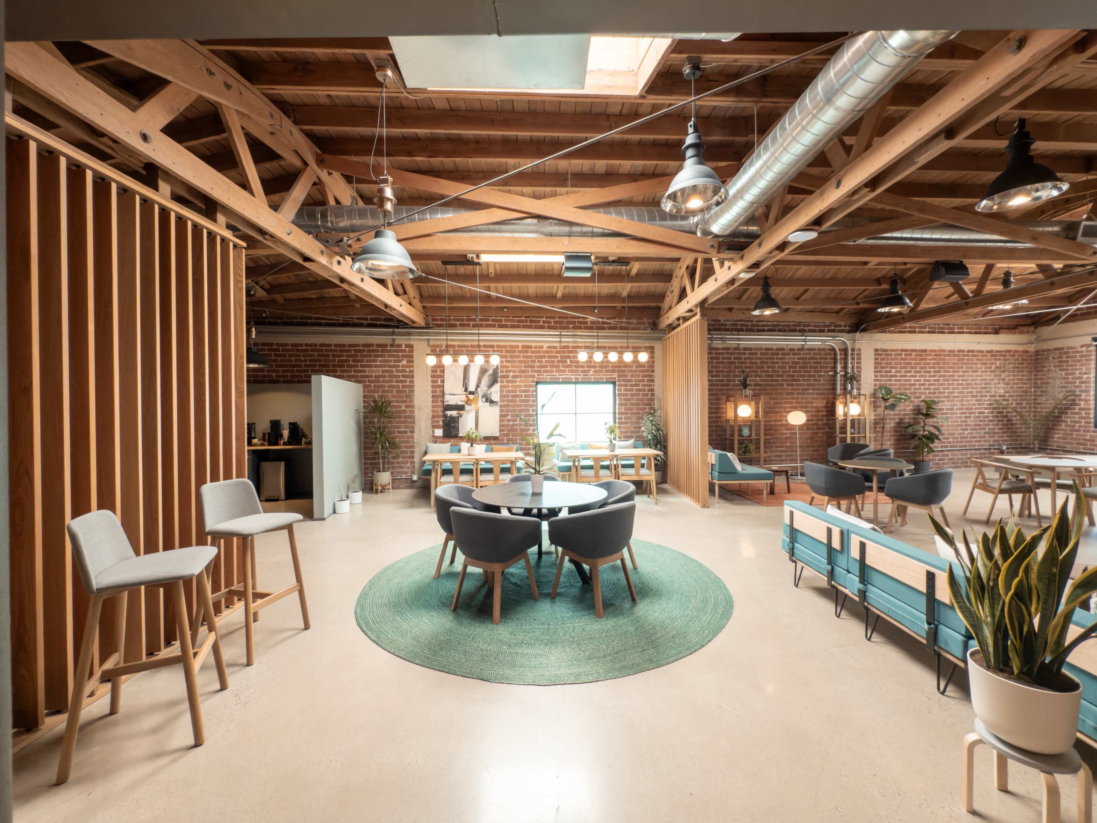 The image shows a spacious, modern workspace with wooden beams, a circular area rug, several seating arrangements, and large windows that allow natural light to enter.