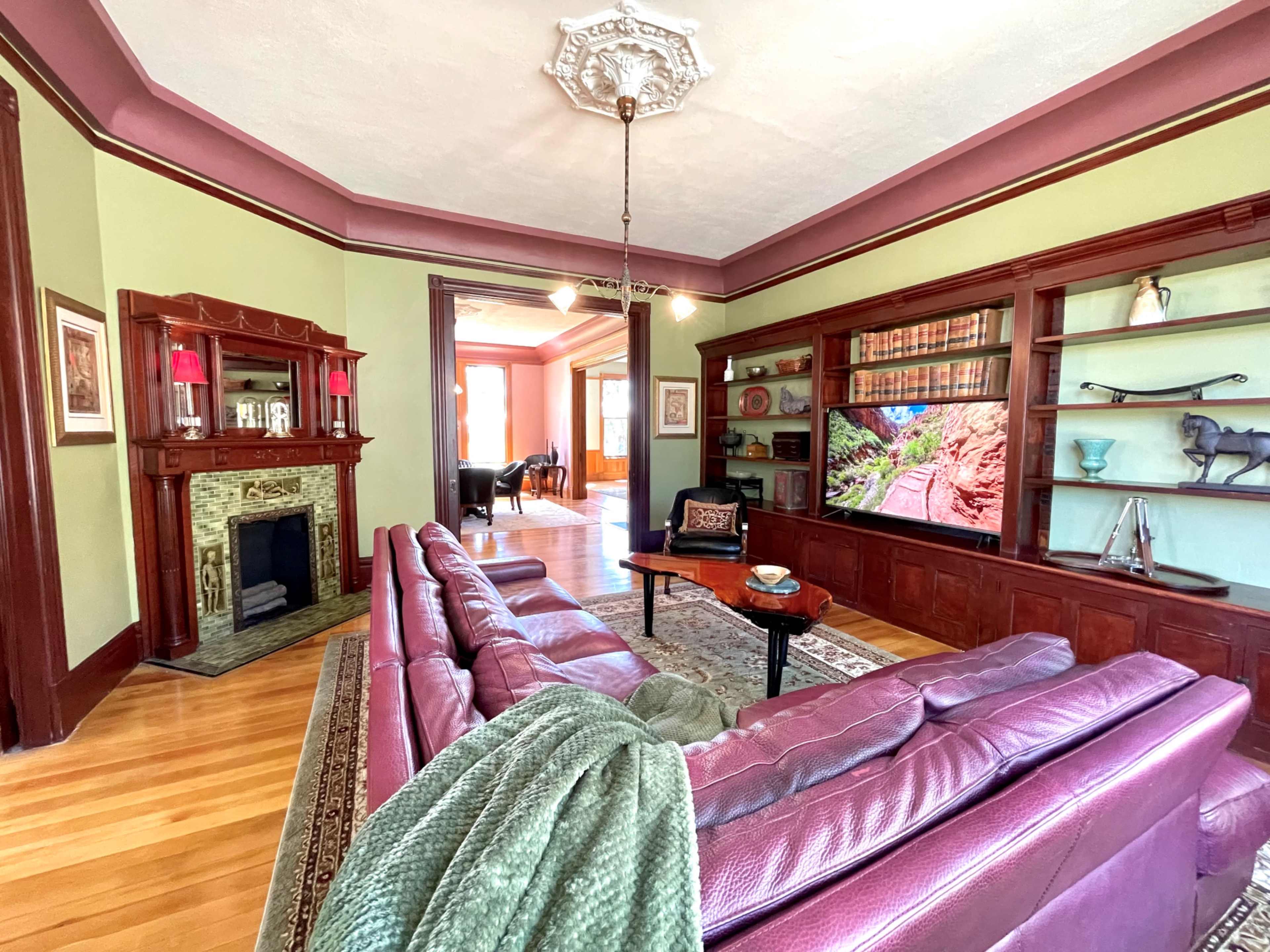 The living room features a burgundy sectional sofa, a small coffee table, a fireplace with an ornate mantel, and built-in shelves displaying decorative items against a light green wall.