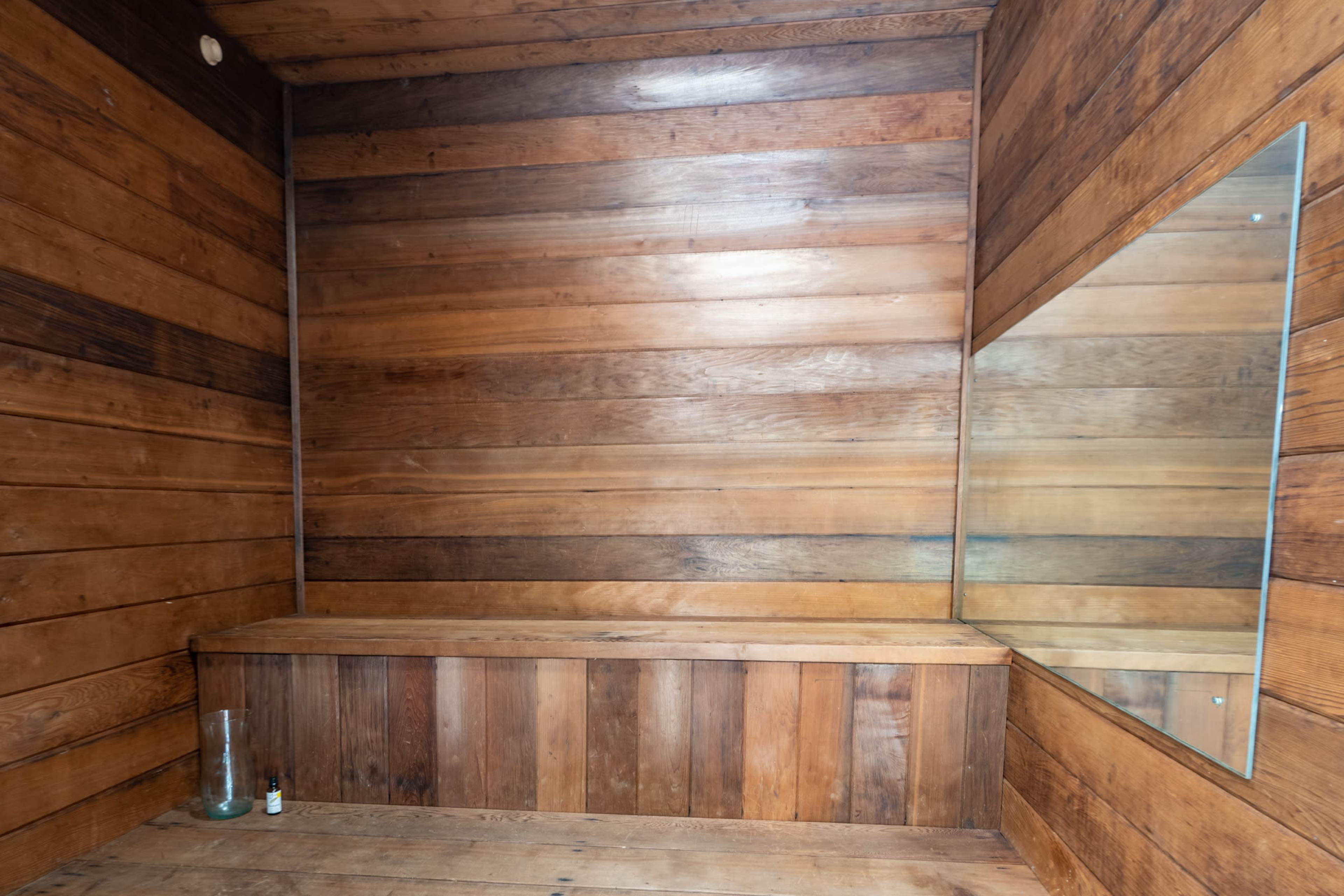 A wooden sauna interior featuring walls and benches made of dark, smooth wood and a large mirror in one corner.
