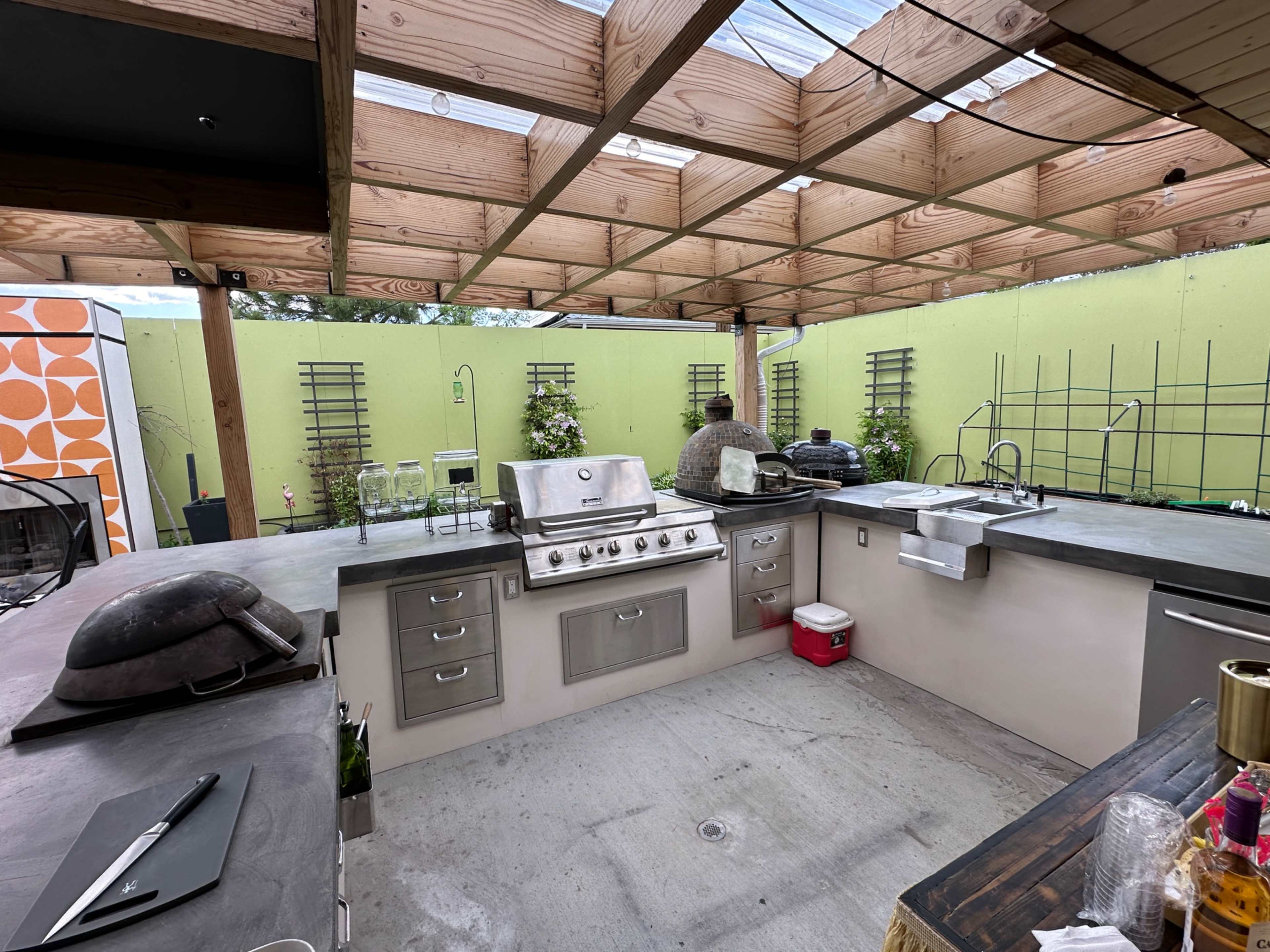 The image shows an outdoor kitchen space featuring a stainless steel grill, countertop areas, and green walls with decorative plants.
