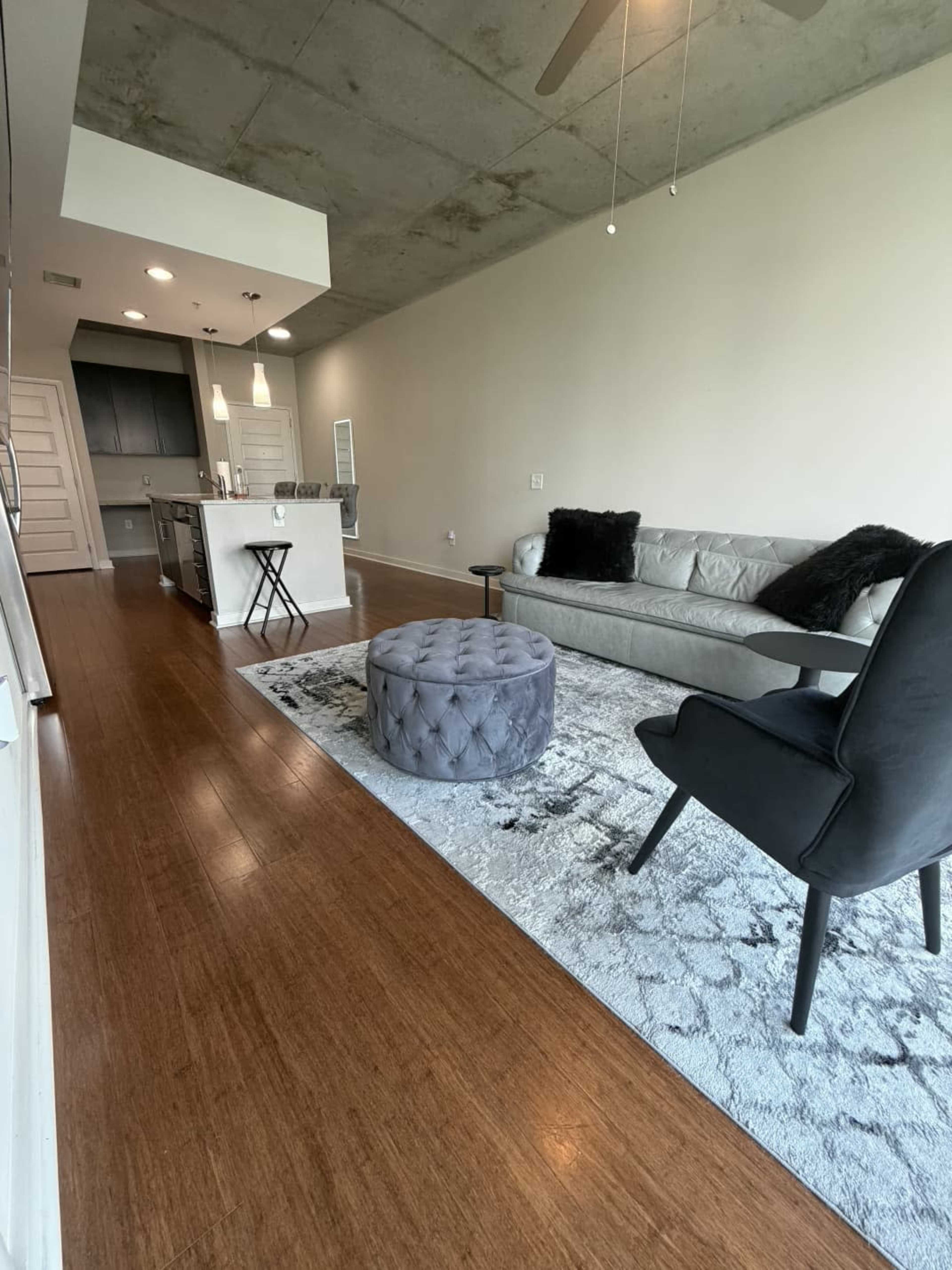 The image shows a modern living room with a gray sofa, a round ottoman, a black chair, and a kitchen area in the background.