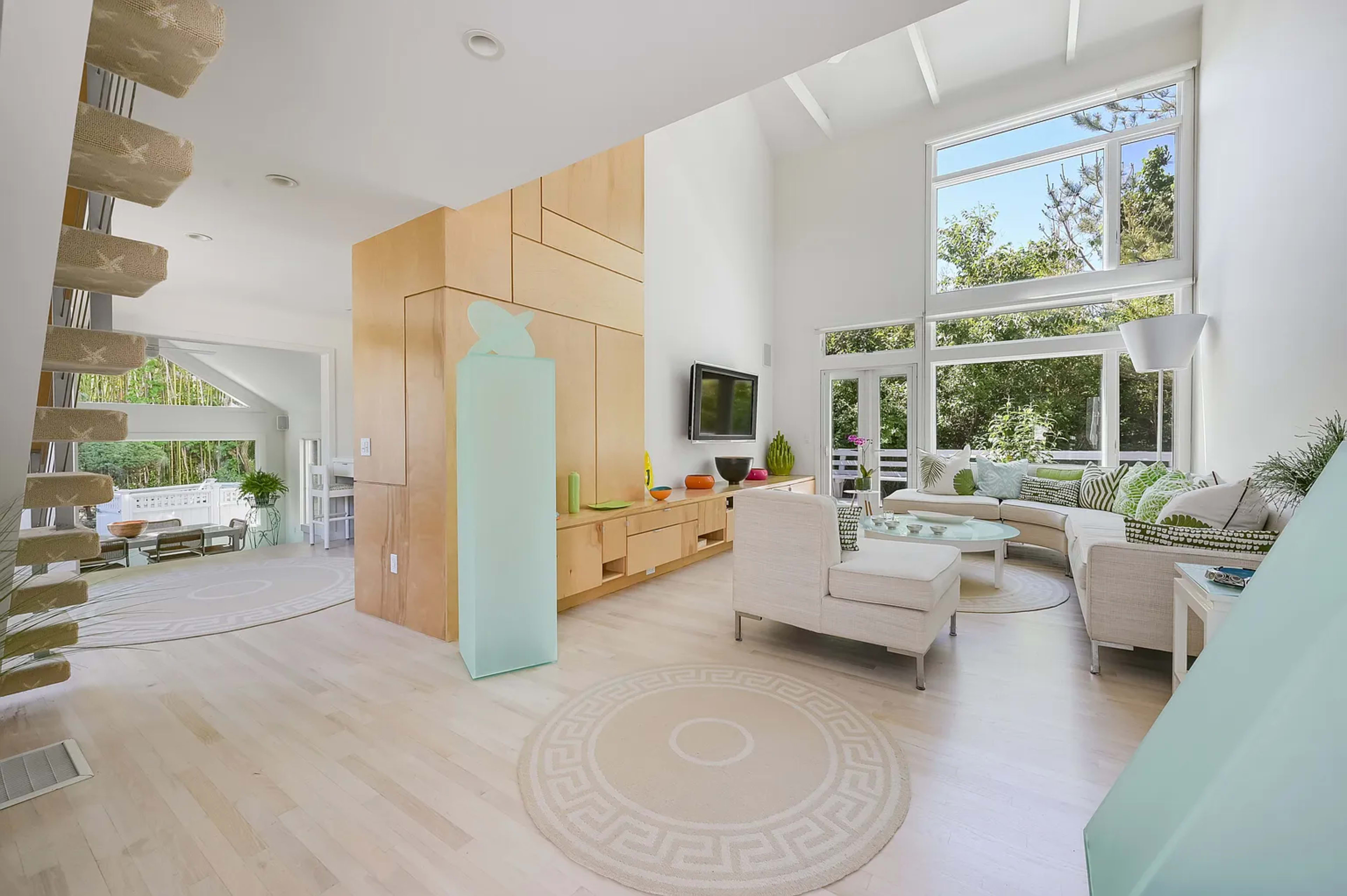 A modern living room features large windows overlooking greenery, a light-colored staircase, and minimalist furniture arranged around a circular area rug.