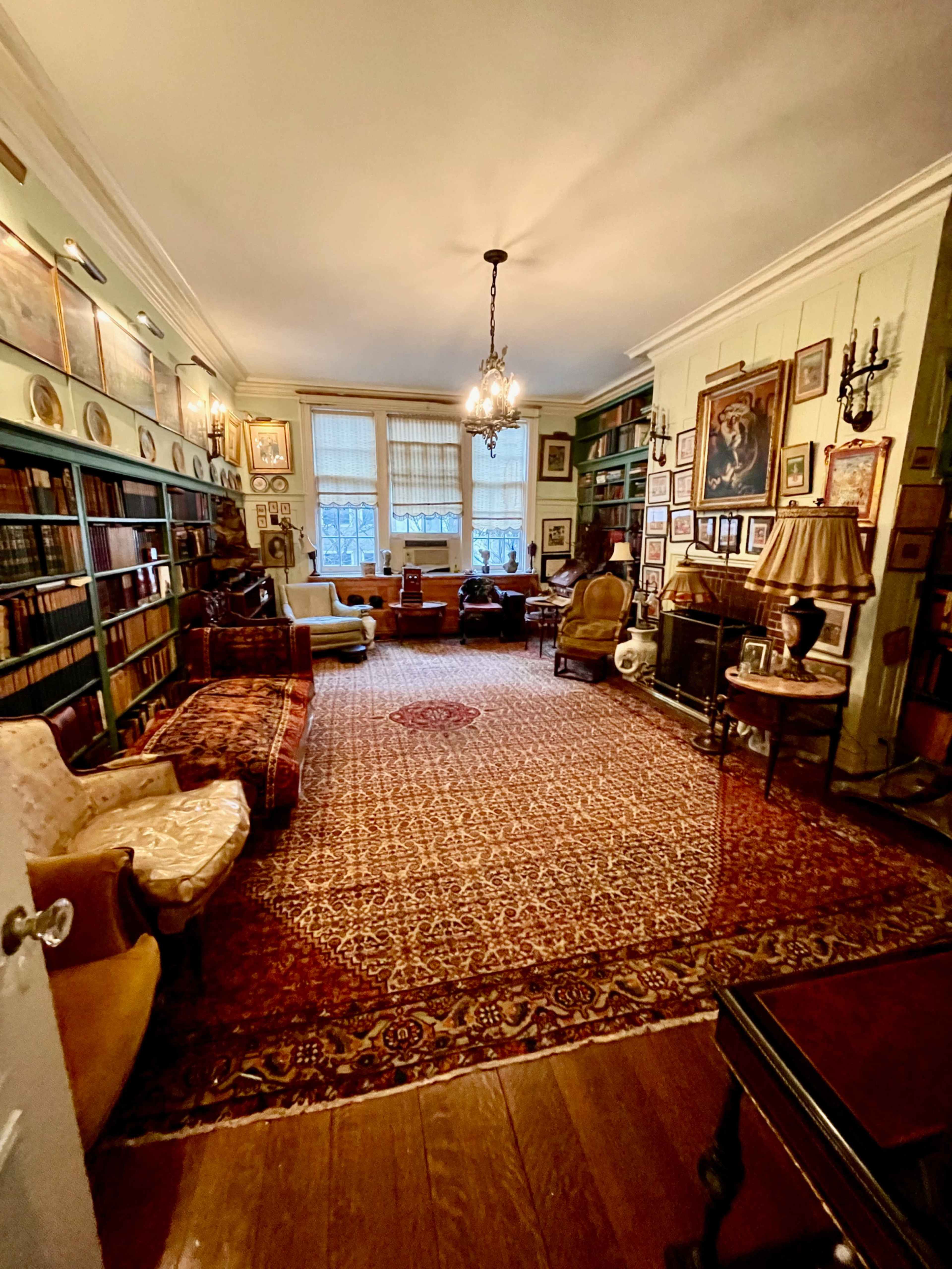 The image shows a spacious, well-furnished library with bookshelves lining the walls, a Persian rug on the floor, and several seating areas.