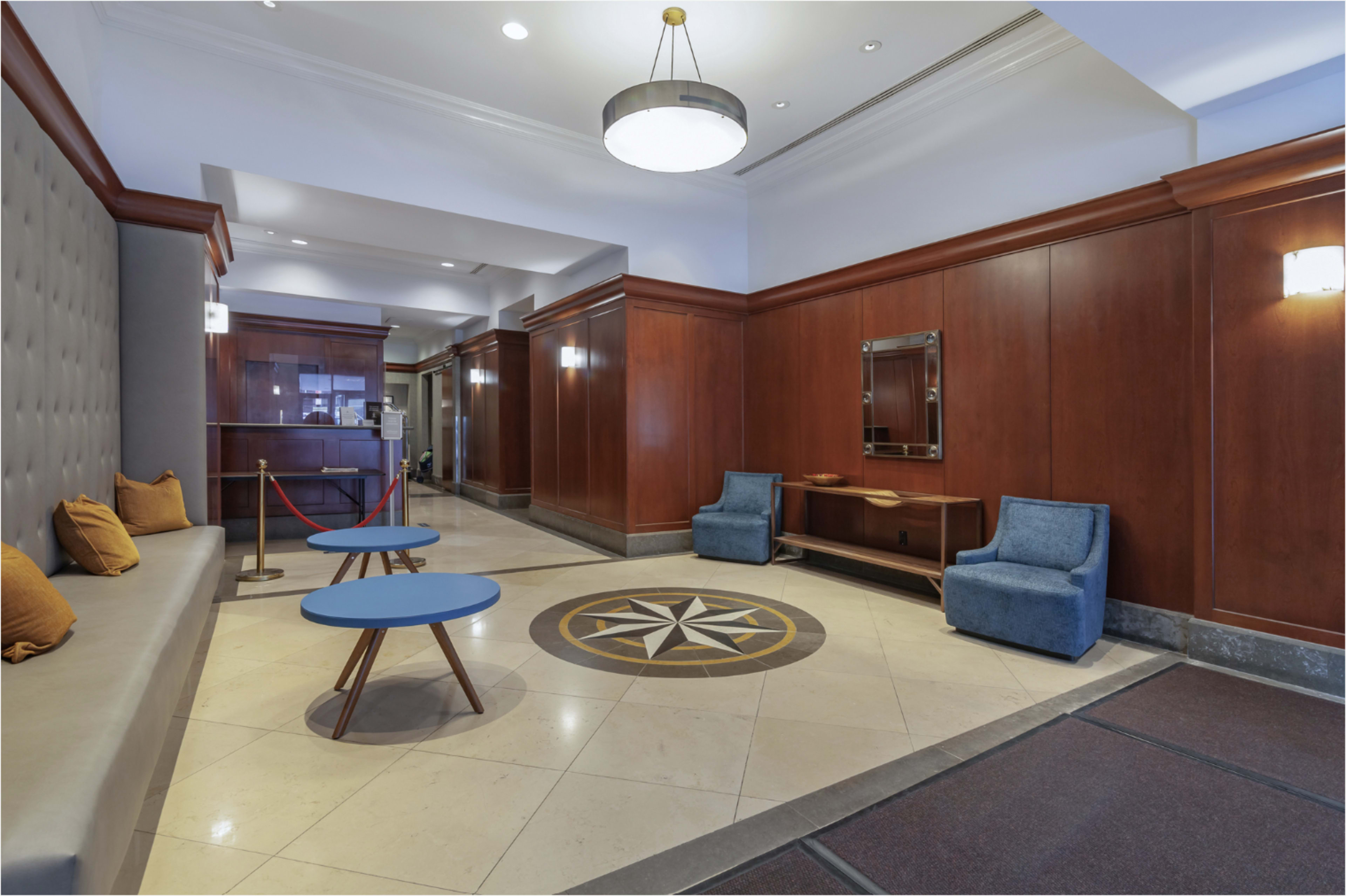 The image shows a lobby area with beige and brown walls, featuring two blue tables, two upholstered chairs, and a star-shaped design in the floor.