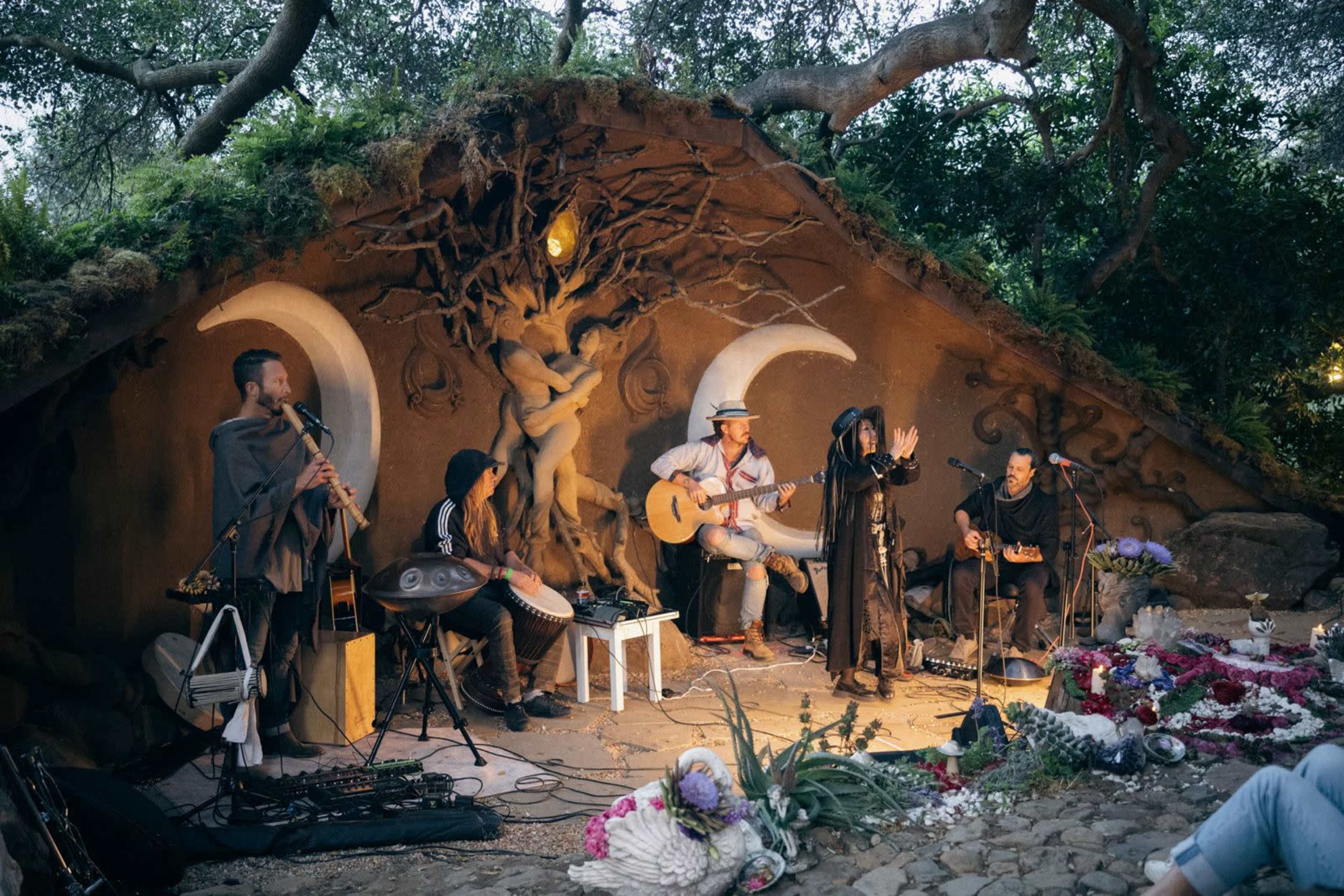 A group of musicians performs in an outdoor setting with a textured wall featuring artistic moon designs and lush greenery surrounding them.
