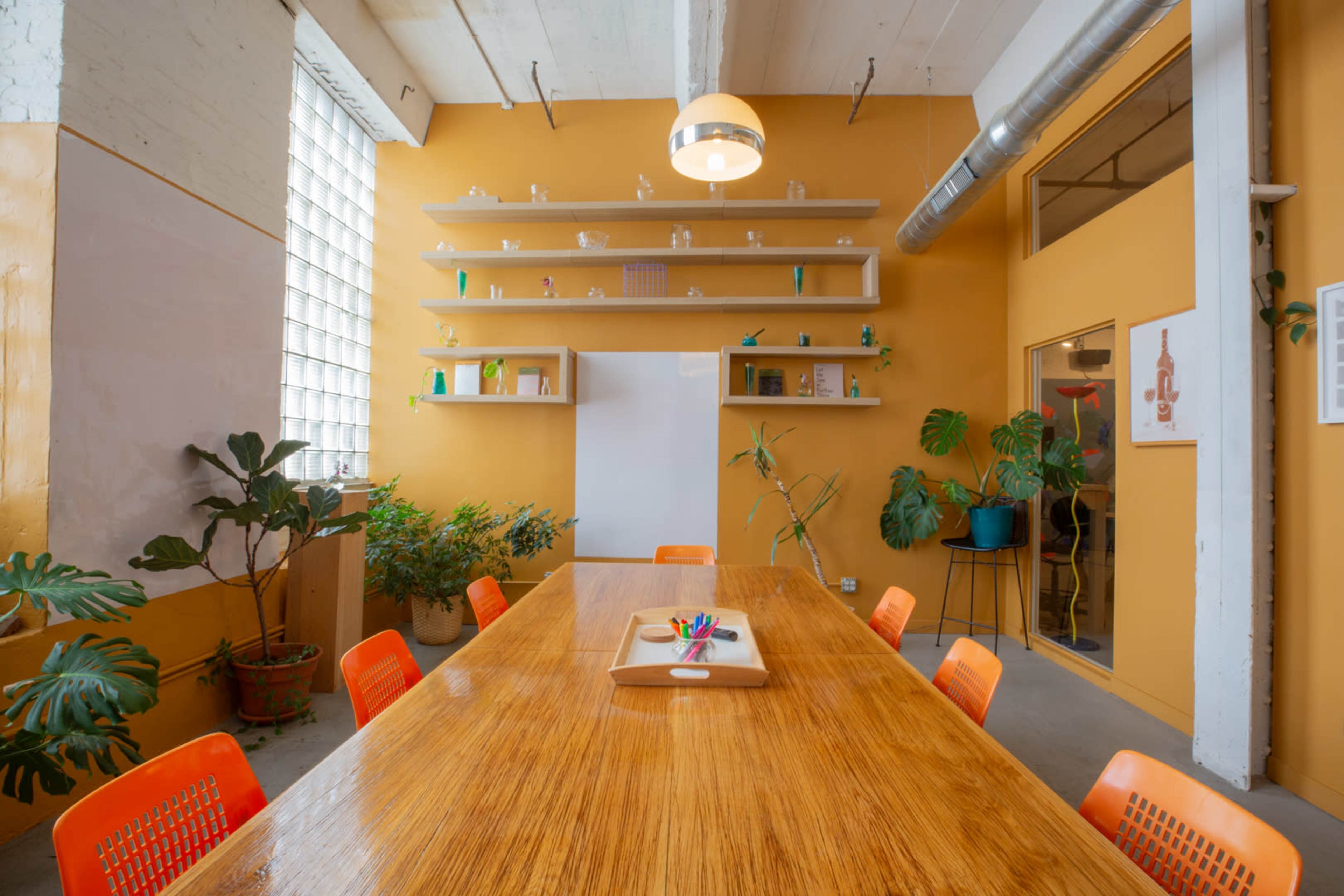 A wooden conference table surrounded by orange chairs is set in a brightly colored meeting room with plants and shelves displaying various items on the walls.