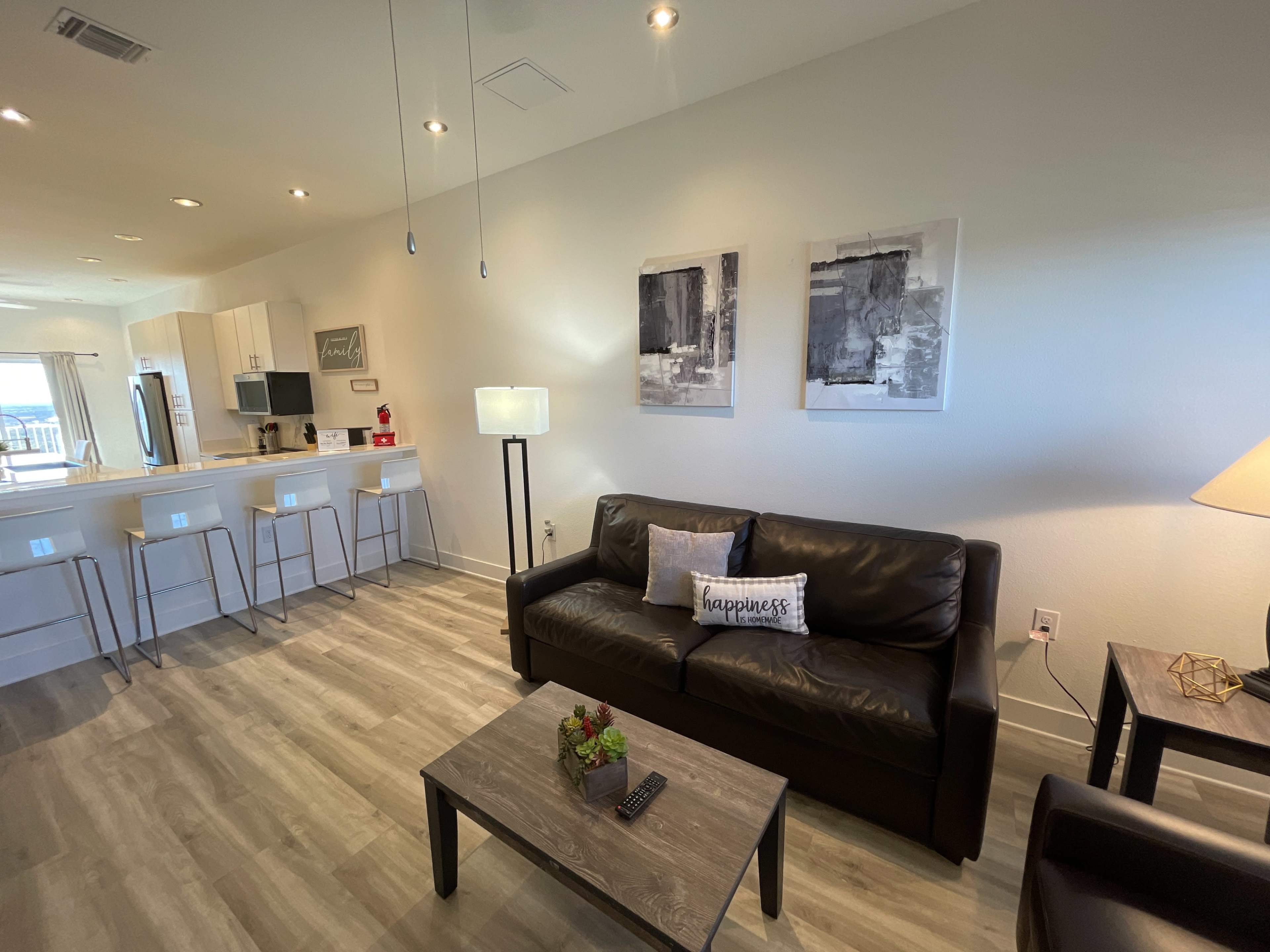 The living area features a black couch and a coffee table, with a lamp and two abstract art pieces on the walls, overlooking a kitchen with bar seating.