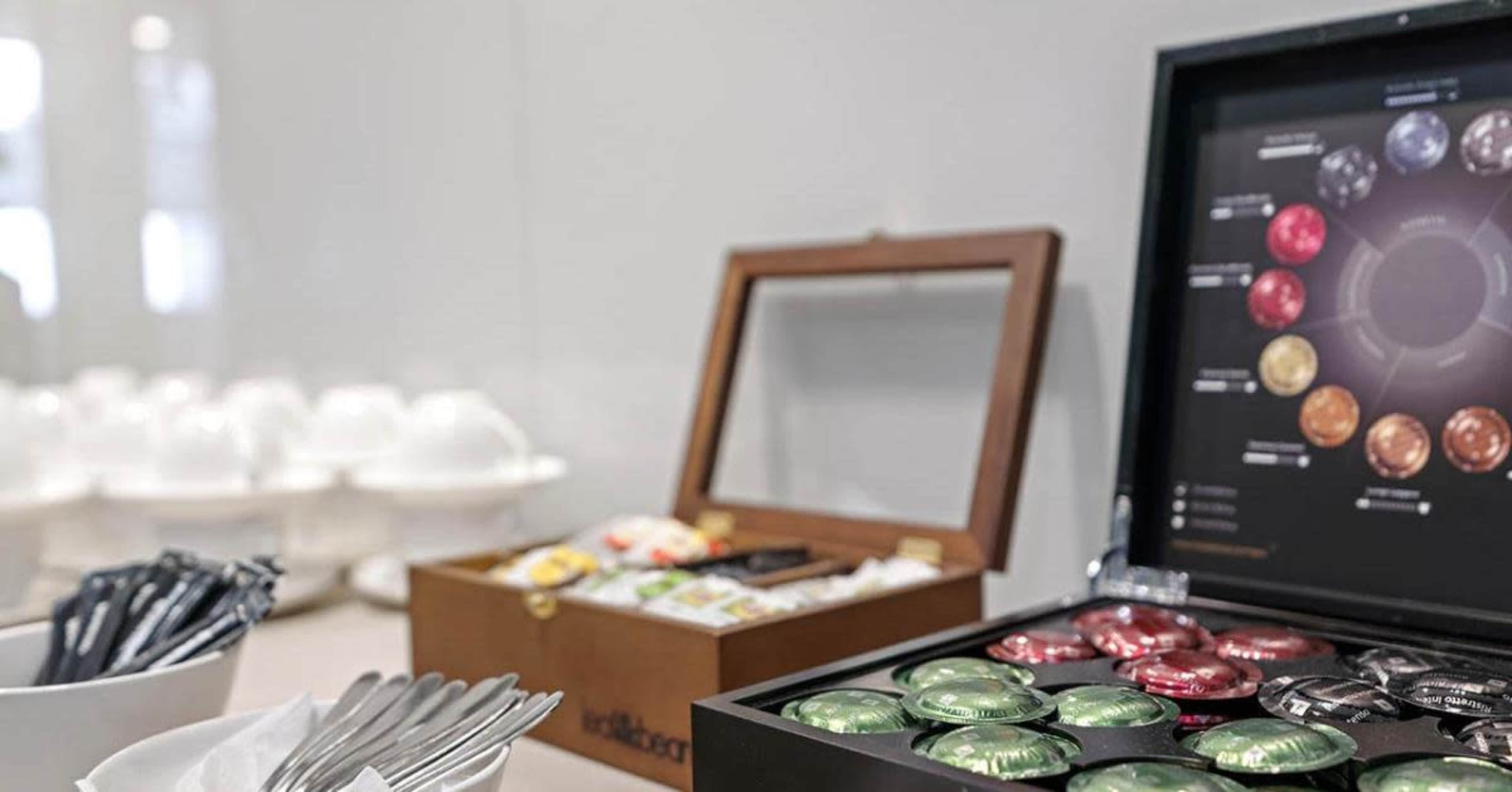 A display of coffee and tea pods in a wooden box alongside utensils and a set of white cups in the background.