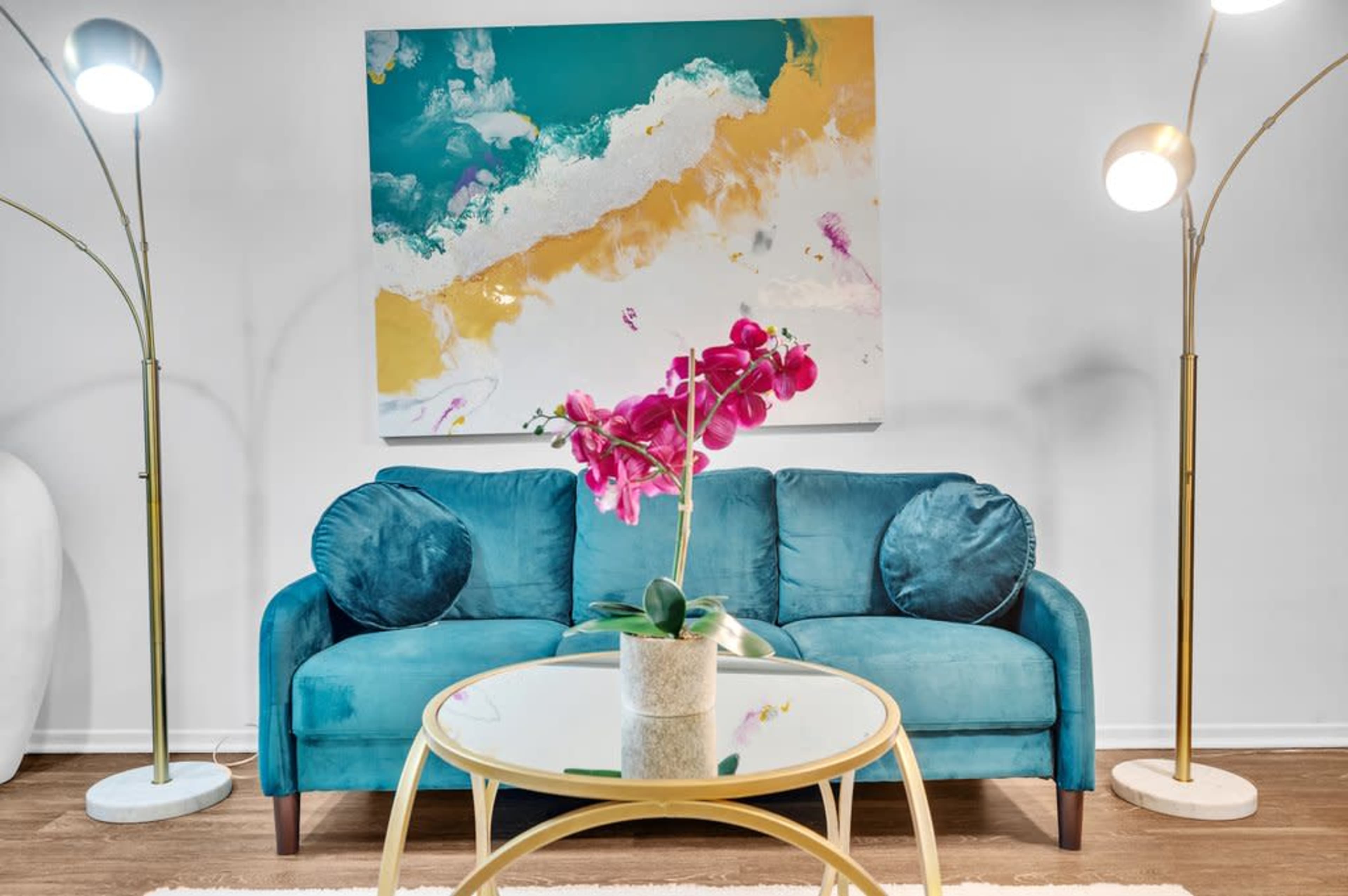 A teal couch with round cushions sits in front of a colorful abstract painting, accompanied by a glass coffee table featuring a potted orchid and two stylish floor lamps.