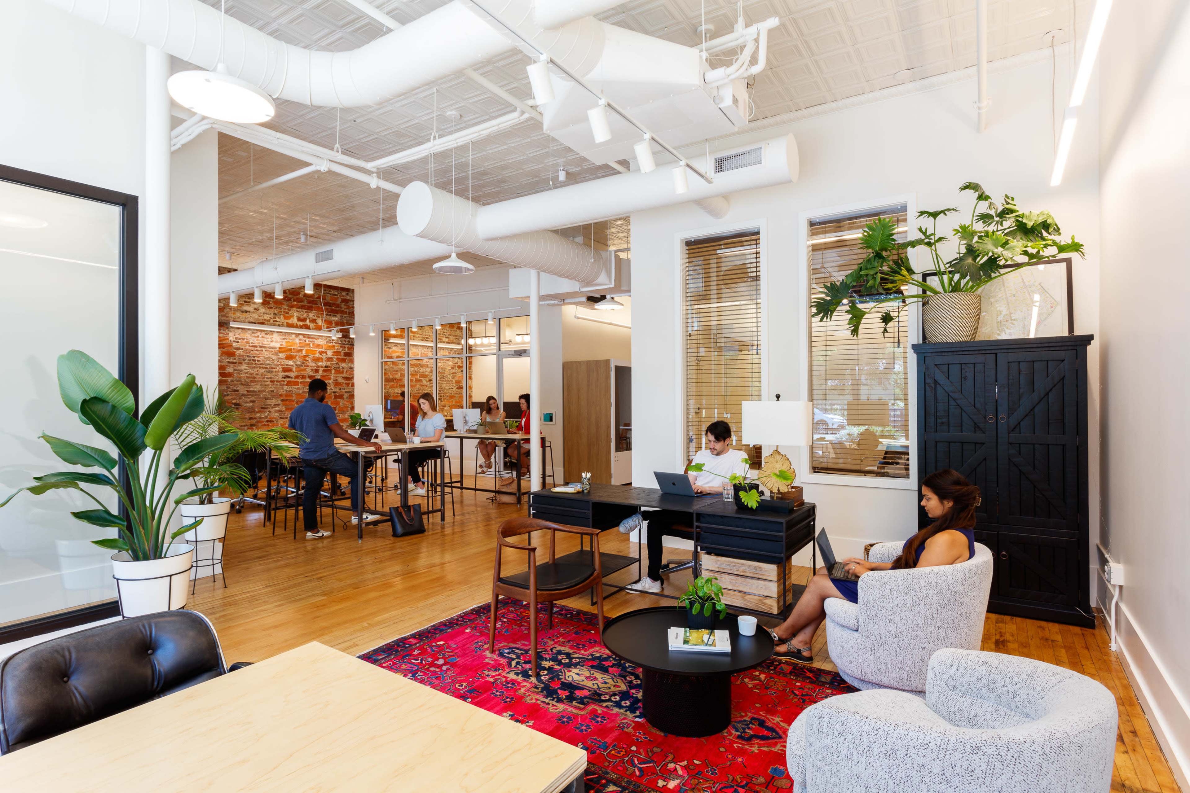 The image shows a modern office space with a mix of workstations and seating areas, featuring exposed brick walls, large windows, and greenery.