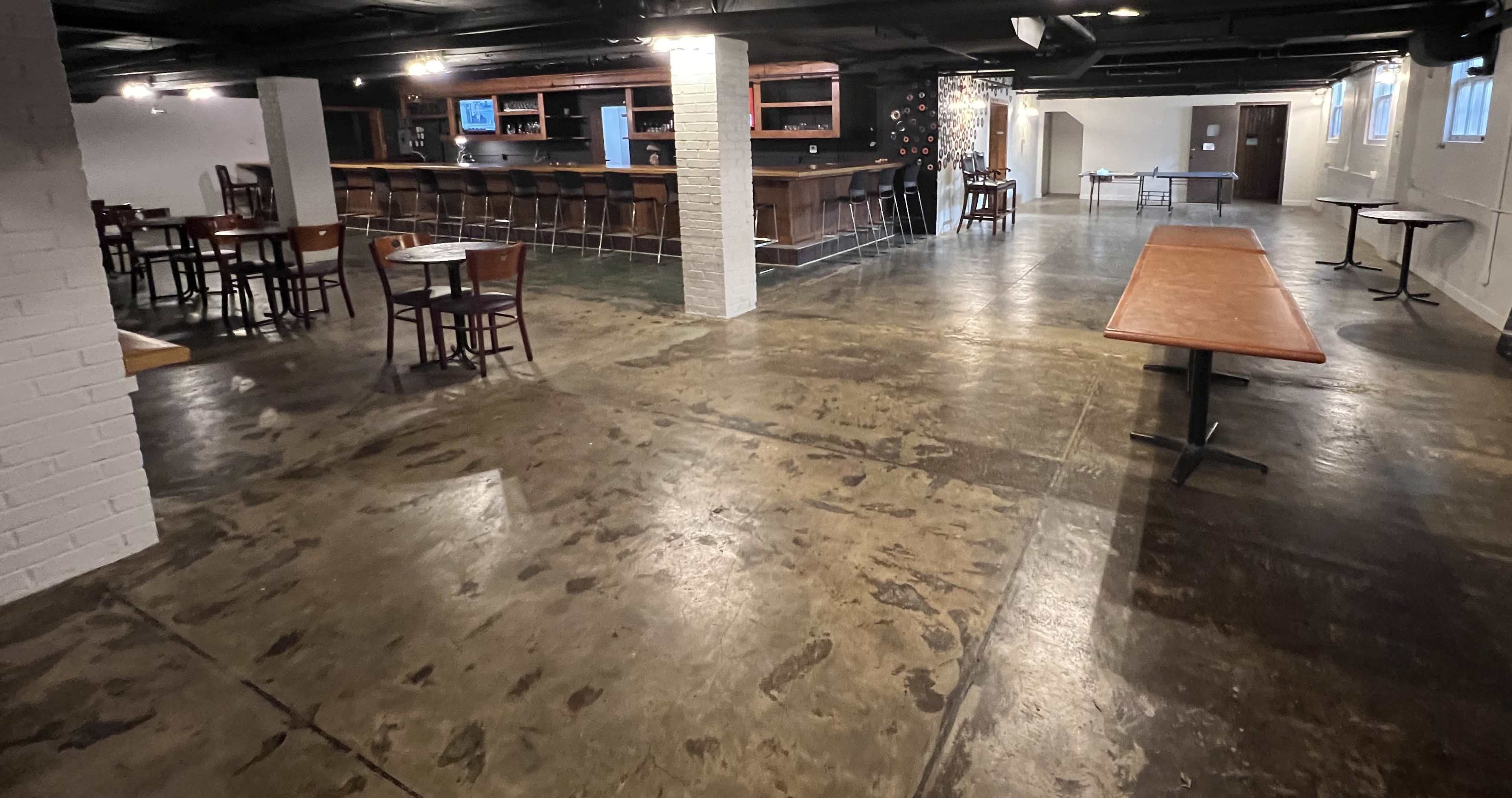 A spacious, empty interior of a bar or lounge area with a long counter, seating arrangements, and polished concrete floors.