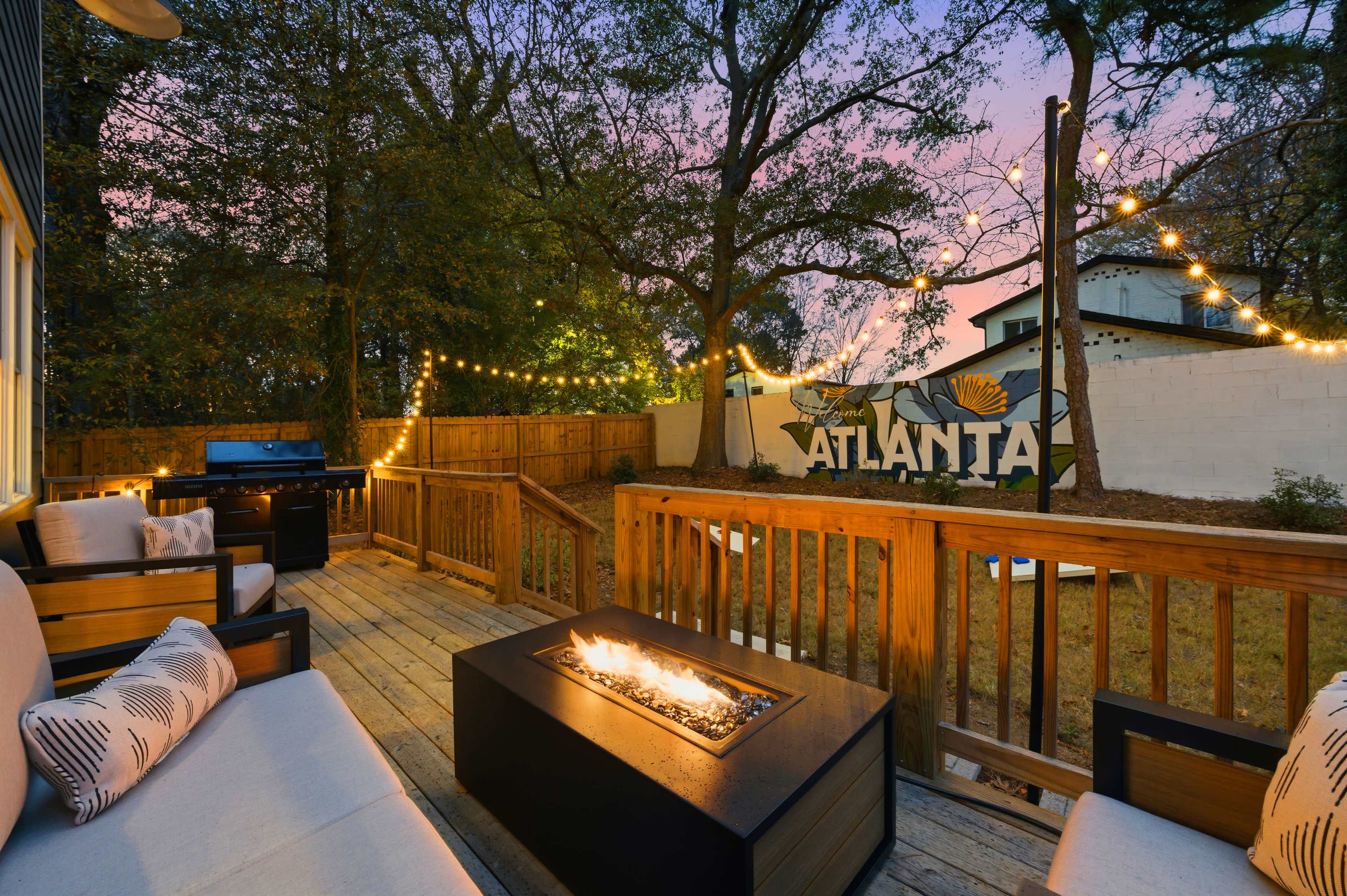 The image shows a wooden deck with modern seating and a fire pit, surrounded by trees and string lights, with a mural that reads "ATLANTA" on a nearby wall.