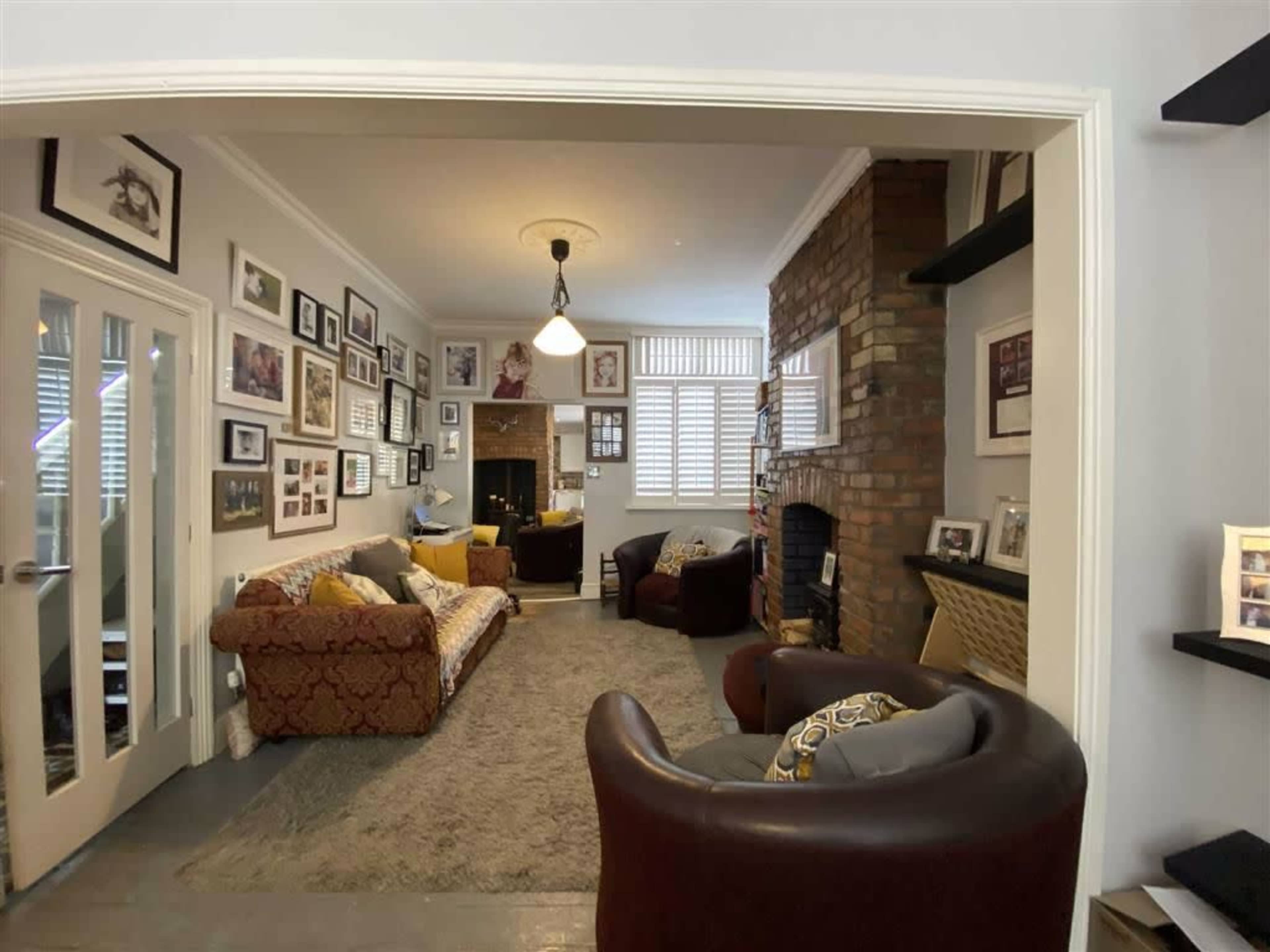 A cozy living room featuring a brick fireplace, framed photographs on the walls, and seating arrangements with a sofa and armchairs.