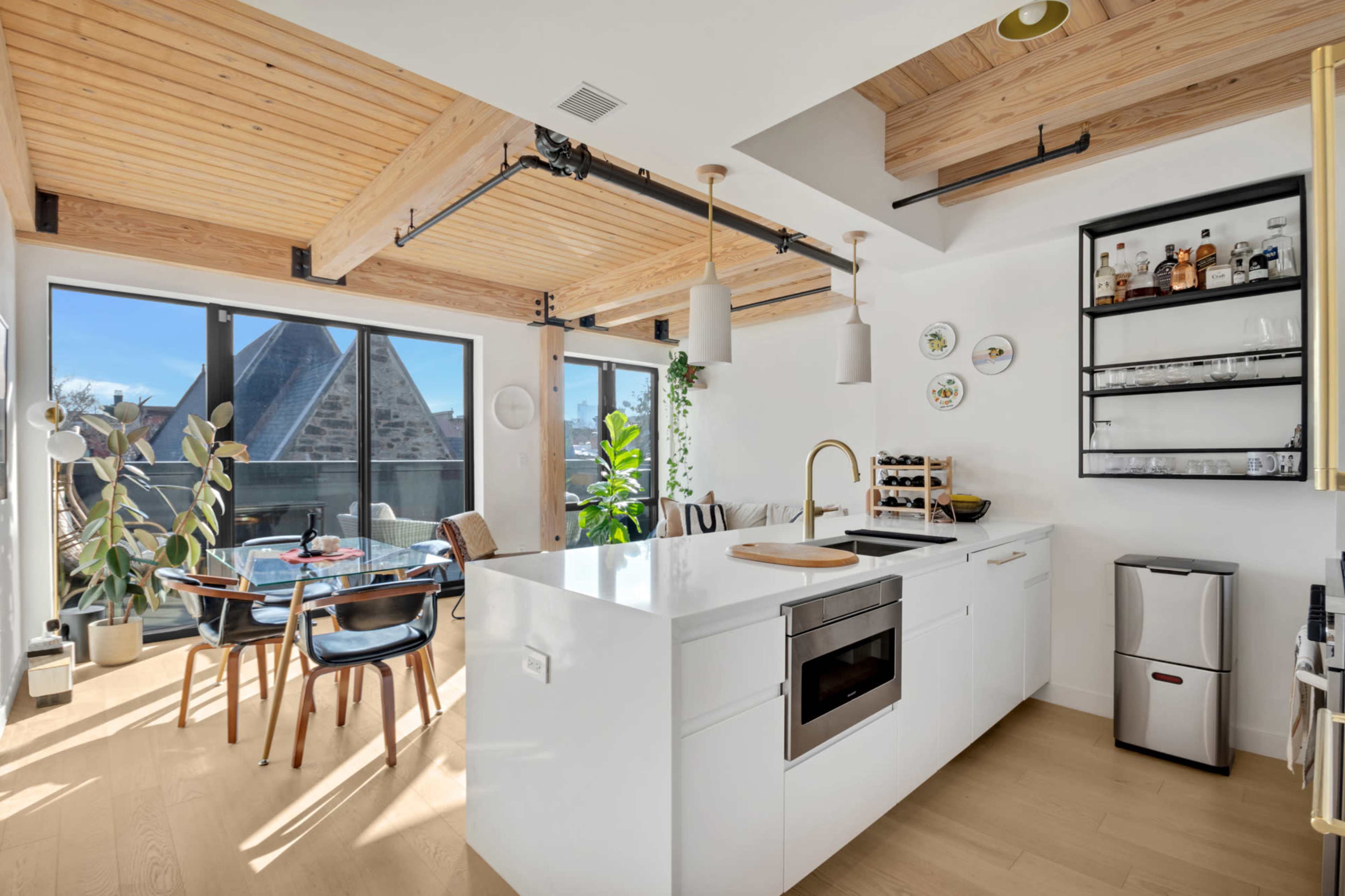The image shows a modern kitchen with a white island, wooden ceiling beams, and large windows that provide natural light, adjacent to a dining area with a table and chairs.