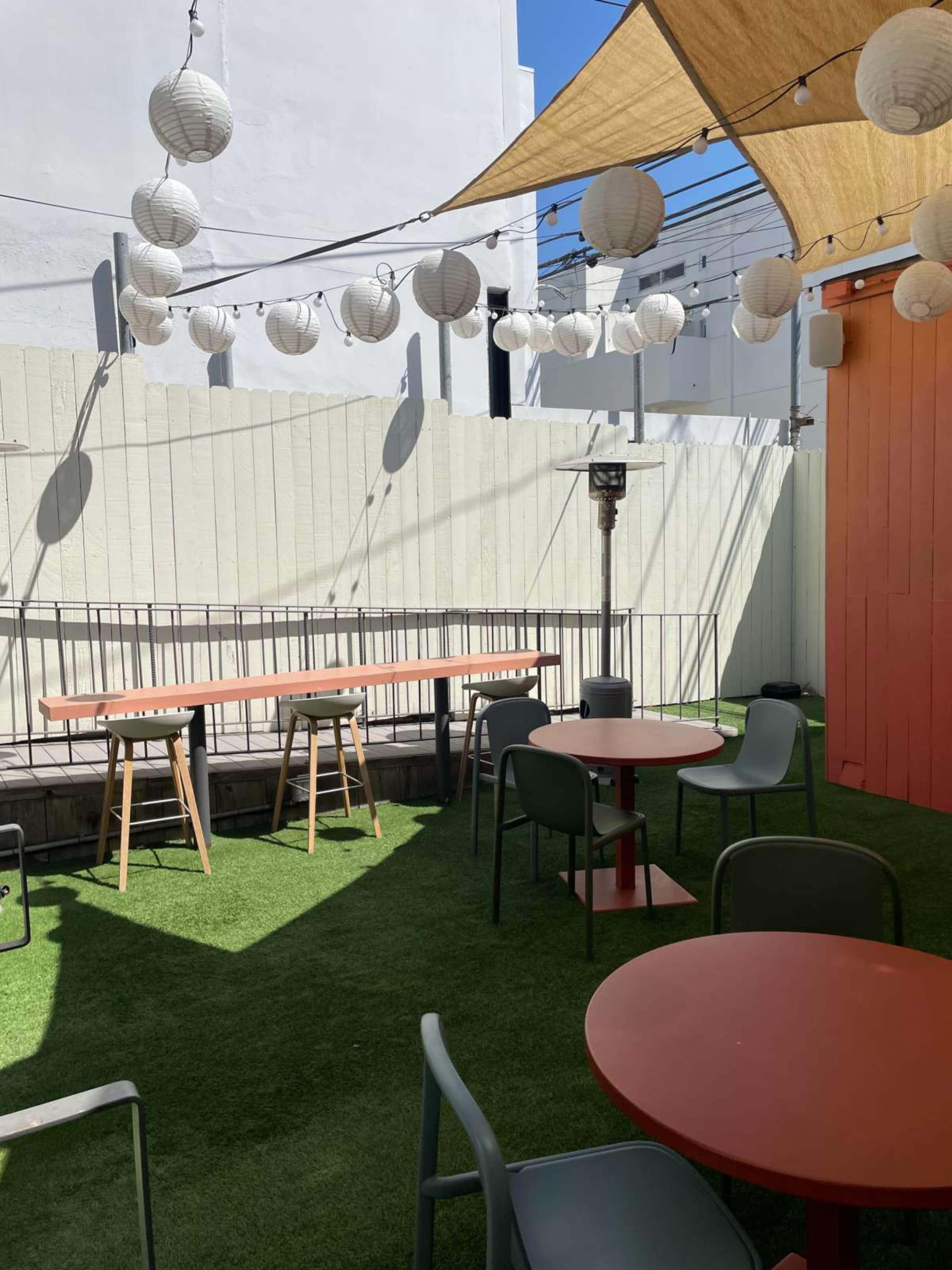 The image shows a sunlit outdoor patio area with green turf, tables, and hanging lanterns.