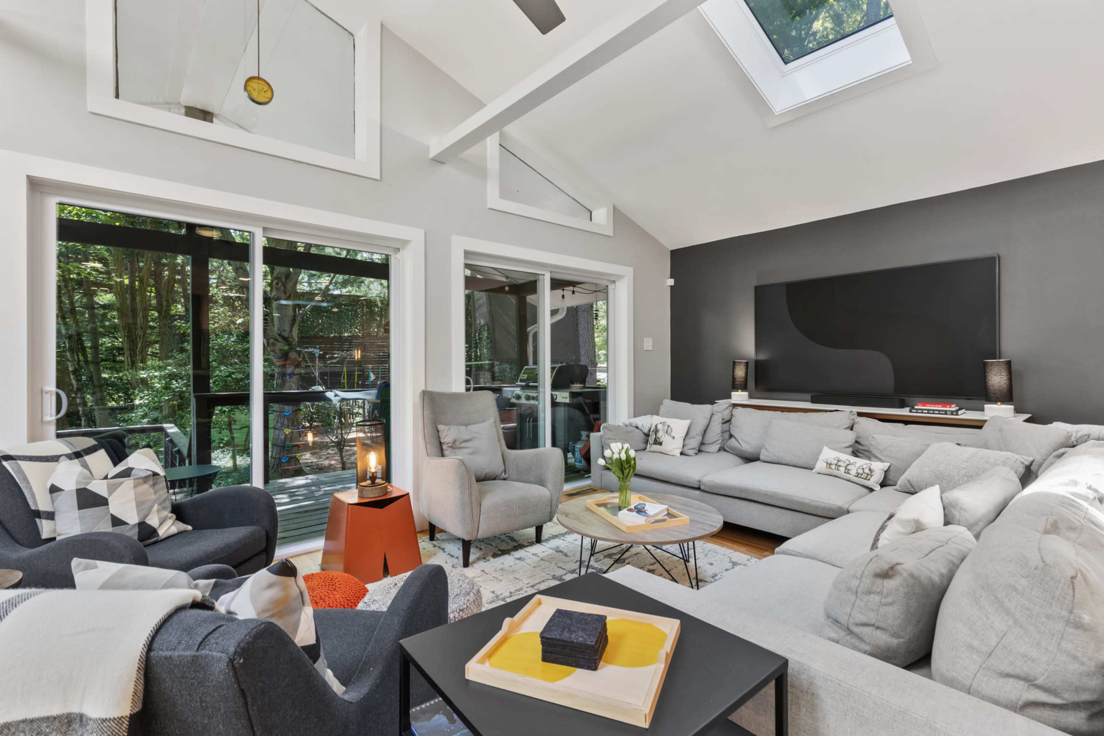 A cozy living room with large windows, modern furniture, and a open layout that connects to a deck overlooking a wooded area.