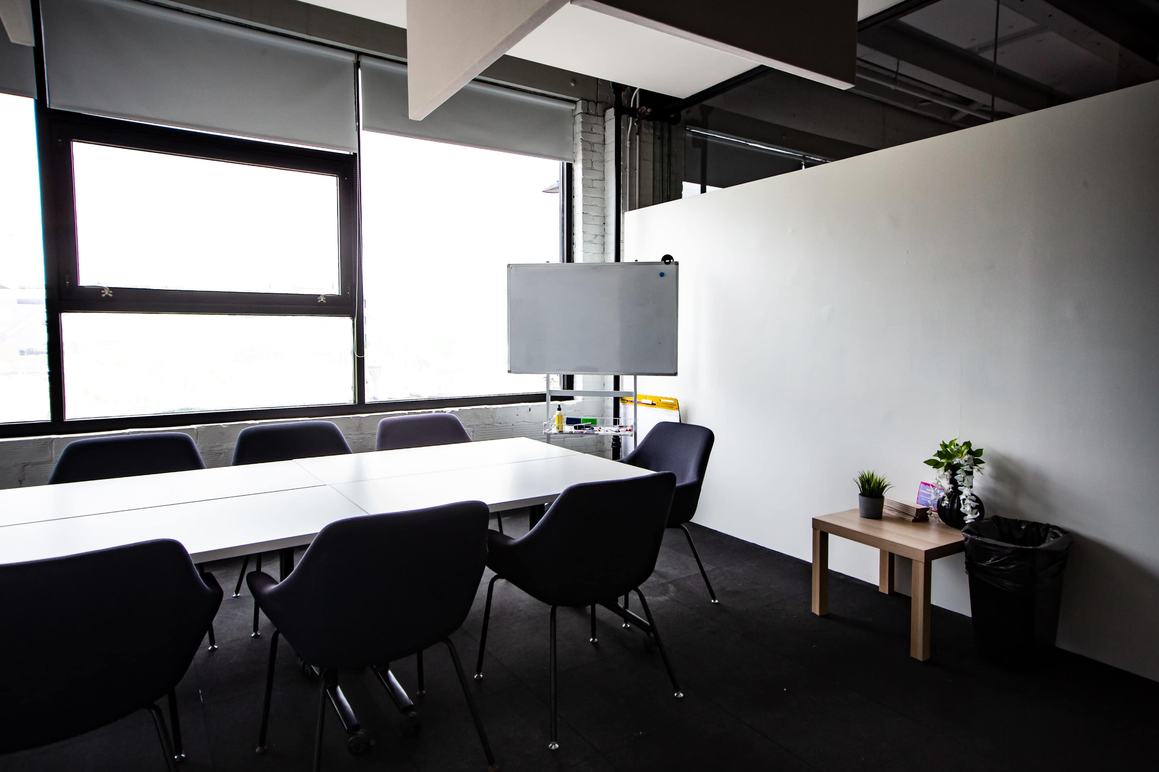 A modern meeting room features a large table surrounded by chairs, with a whiteboard and a small table with a plant near a wall.