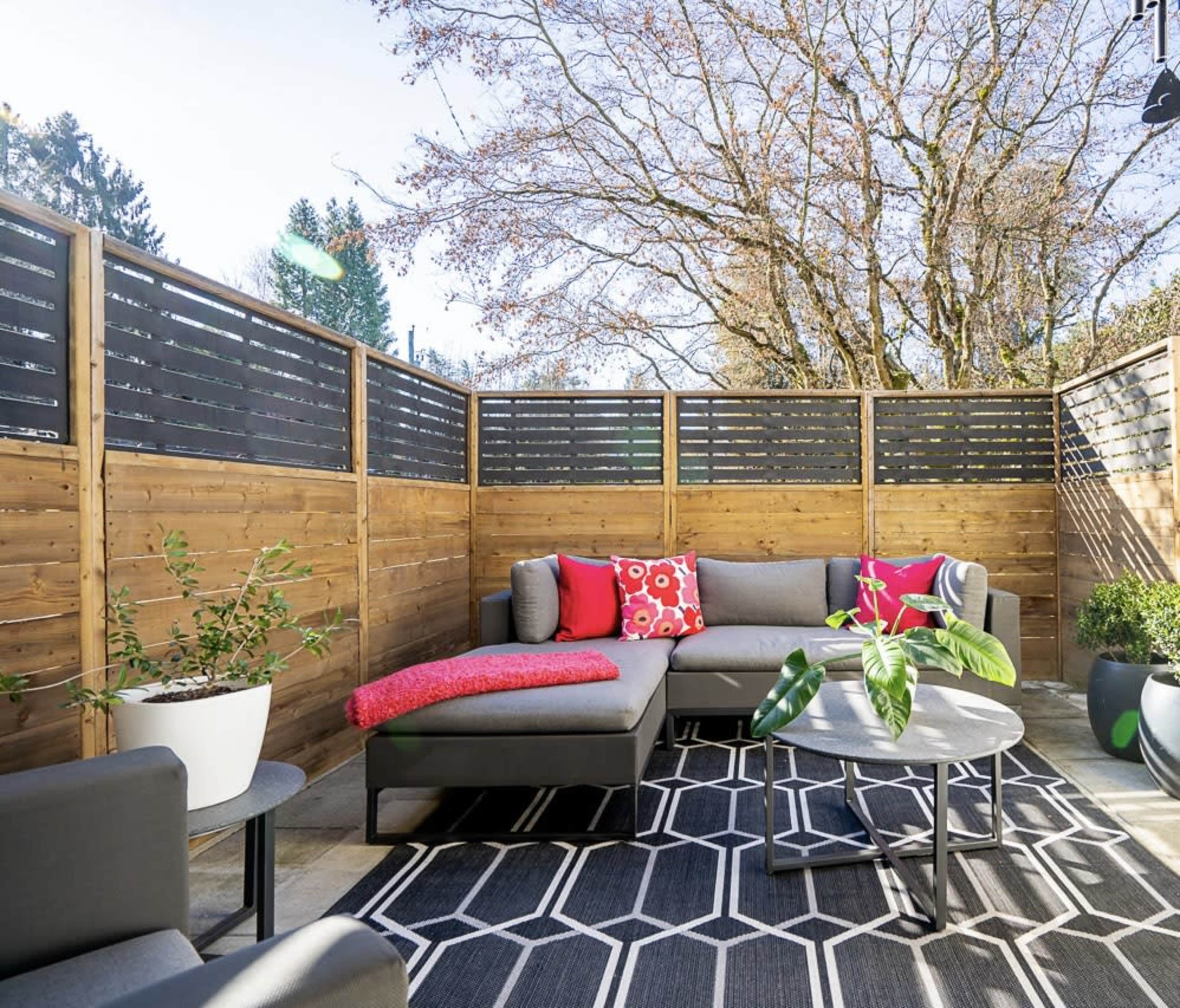 A modern outdoor seating area features a sectional sofa with colorful cushions, a round coffee table, and potted plants, surrounded by wooden fencing and a backdrop of trees.