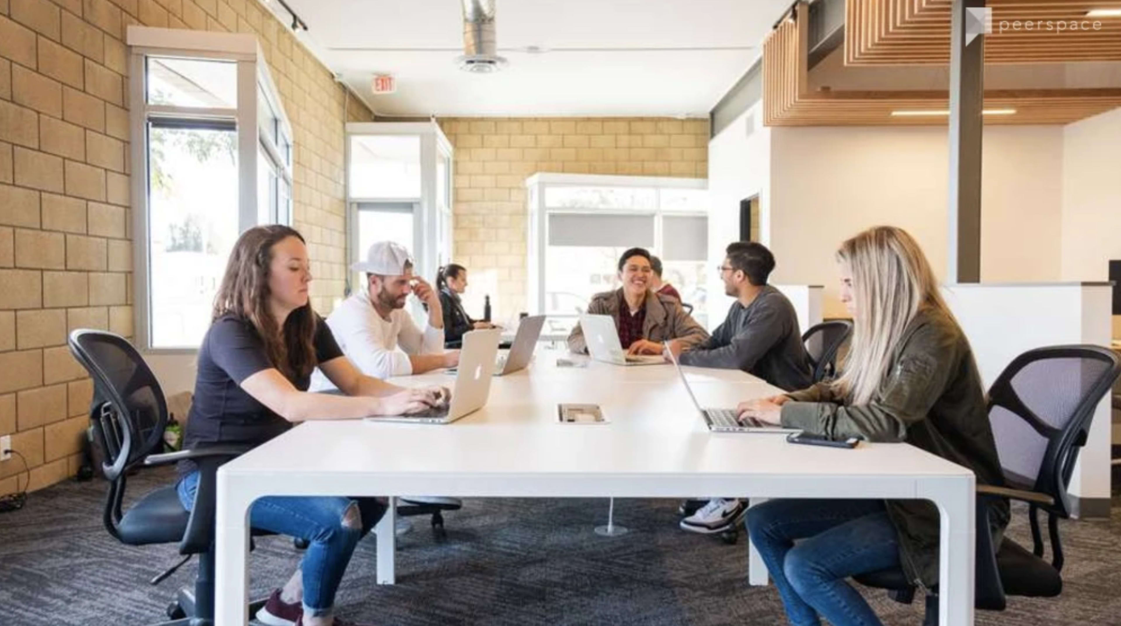 A group of six people work on laptops at a long, white table in a modern office space.