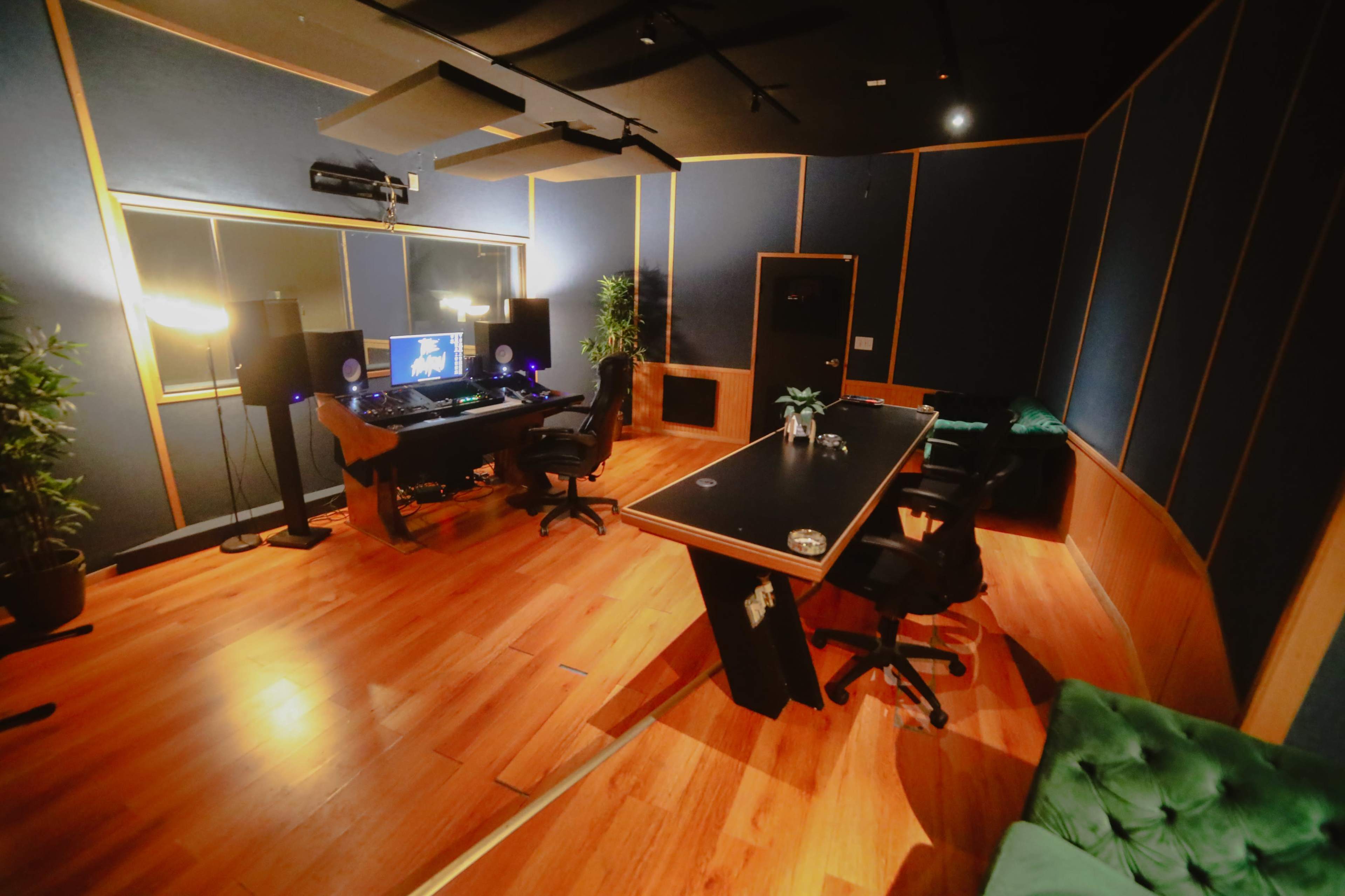 The image depicts a modern music studio featuring a computer workstation, audio equipment, and a large table surrounded by chairs, all set on polished wooden flooring.