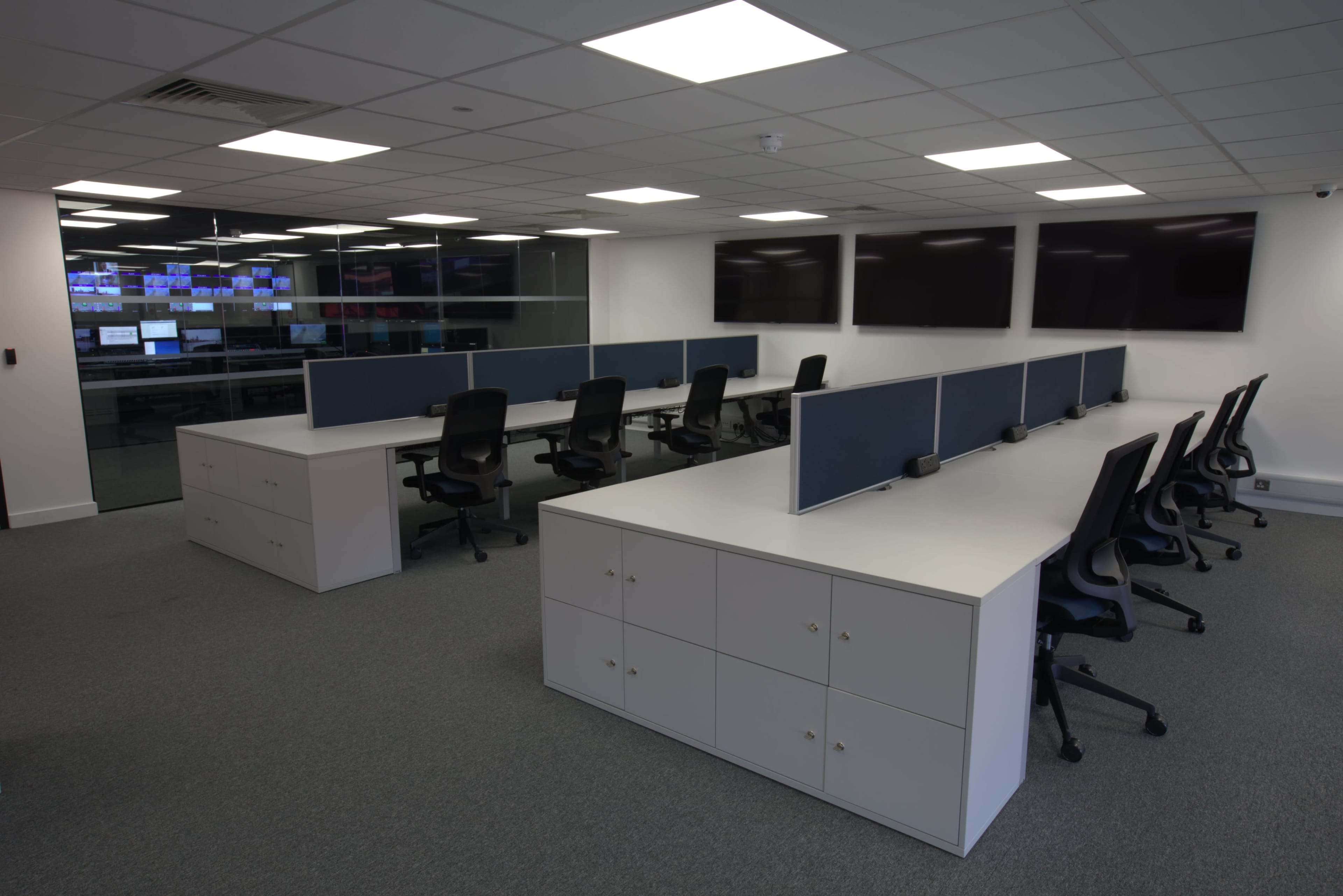 The image shows a modern office space with multiple desks and chairs, organized in a spacious layout, and large screens mounted on the wall.