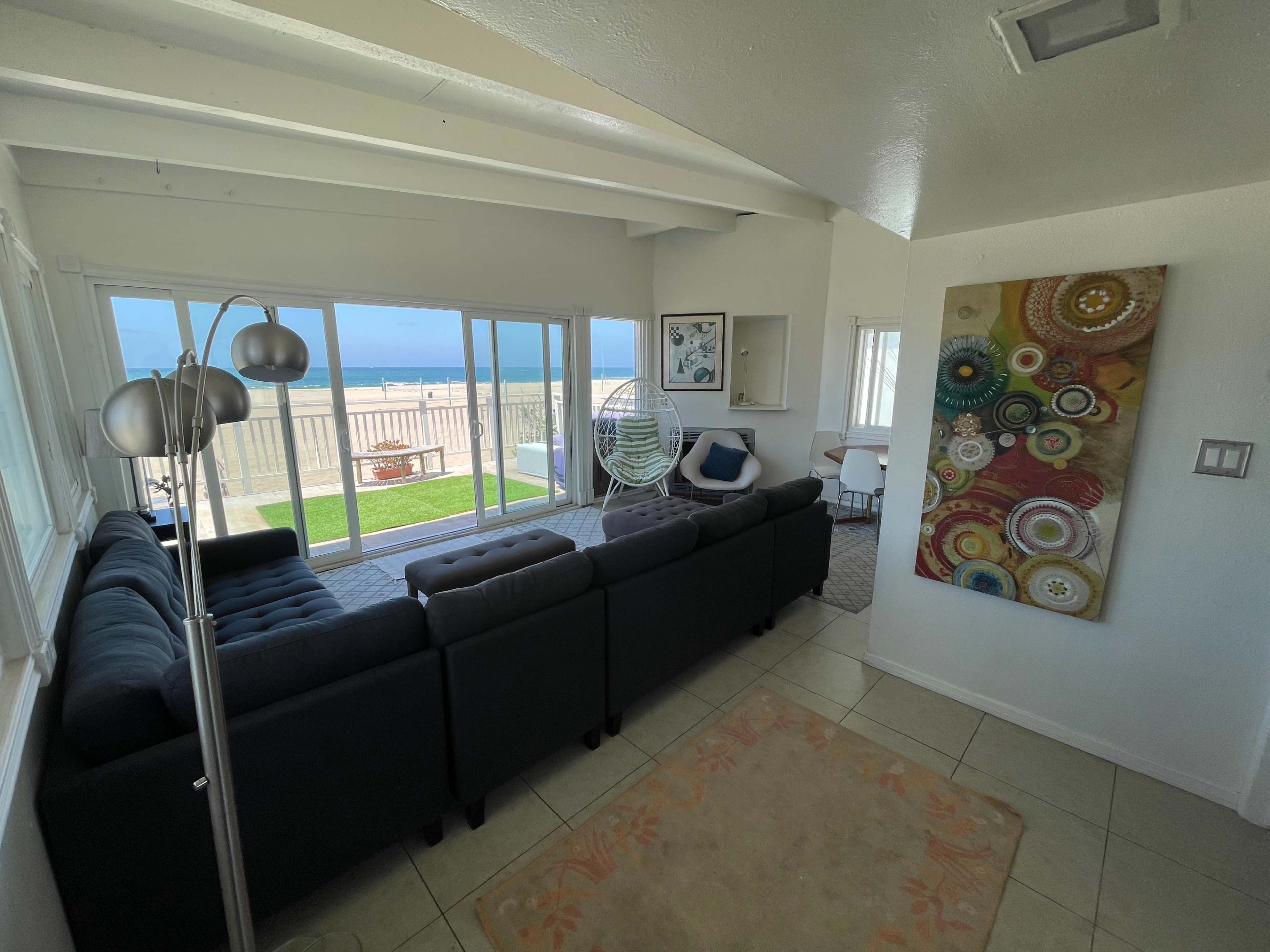 A modern living room features a dark sectional sofa, a colorful wall art piece, and large windows overlooking a grassy outdoor space.
