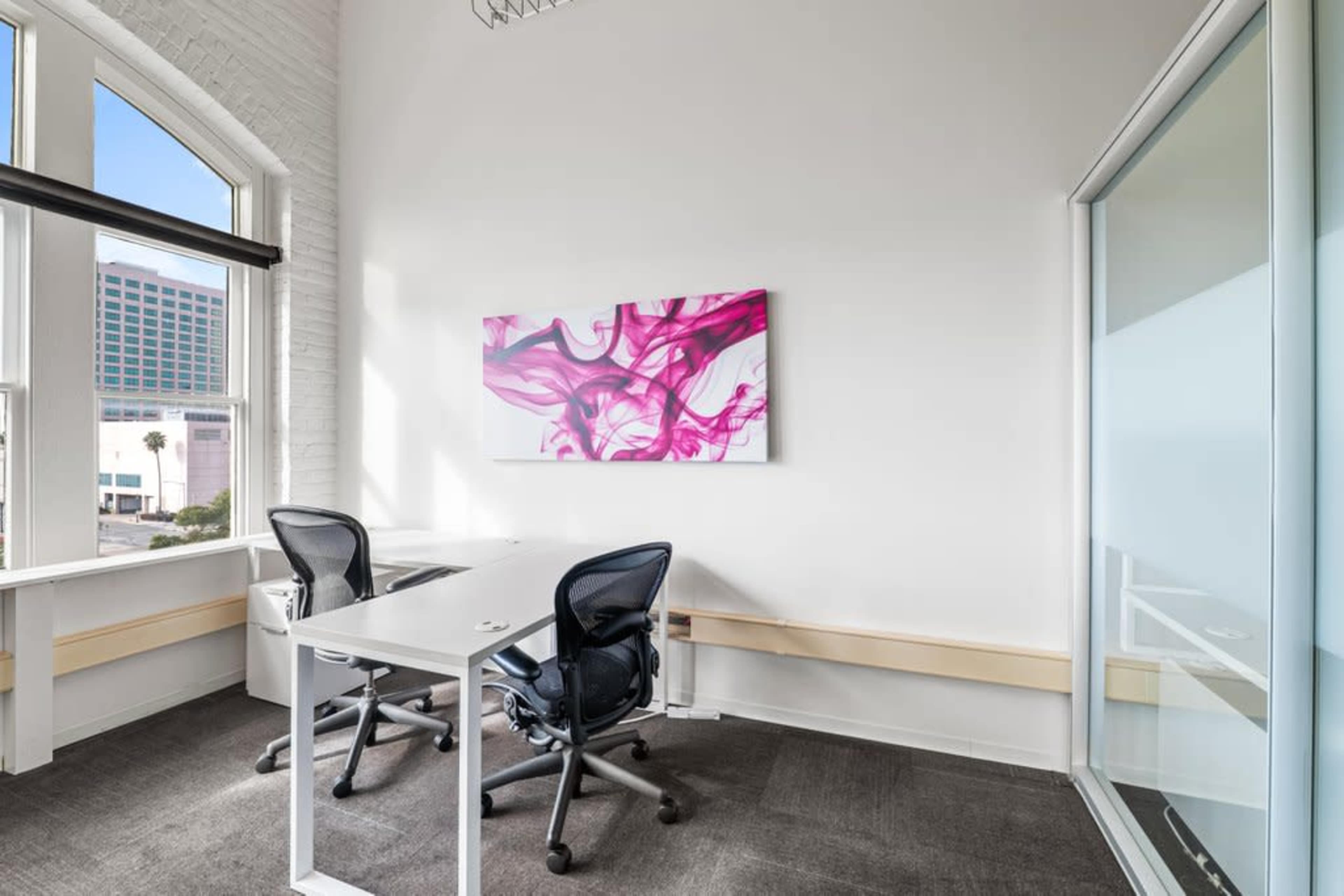 A modern office space features two desks with chairs, a large vibrant pink artwork on the wall, and large windows allowing natural light to enter.