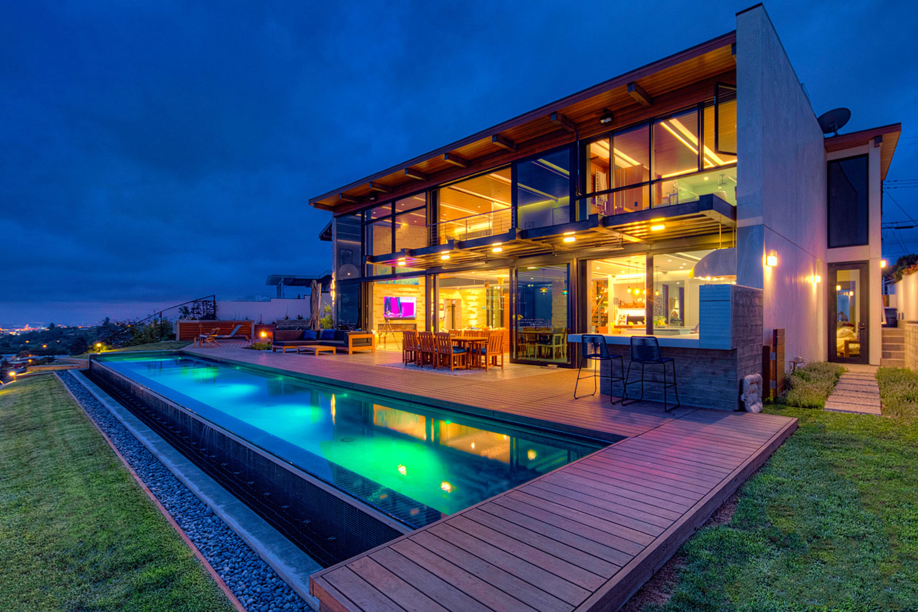 A modern two-story house features large windows and a wooden deck with a swimming pool reflecting the evening sky.