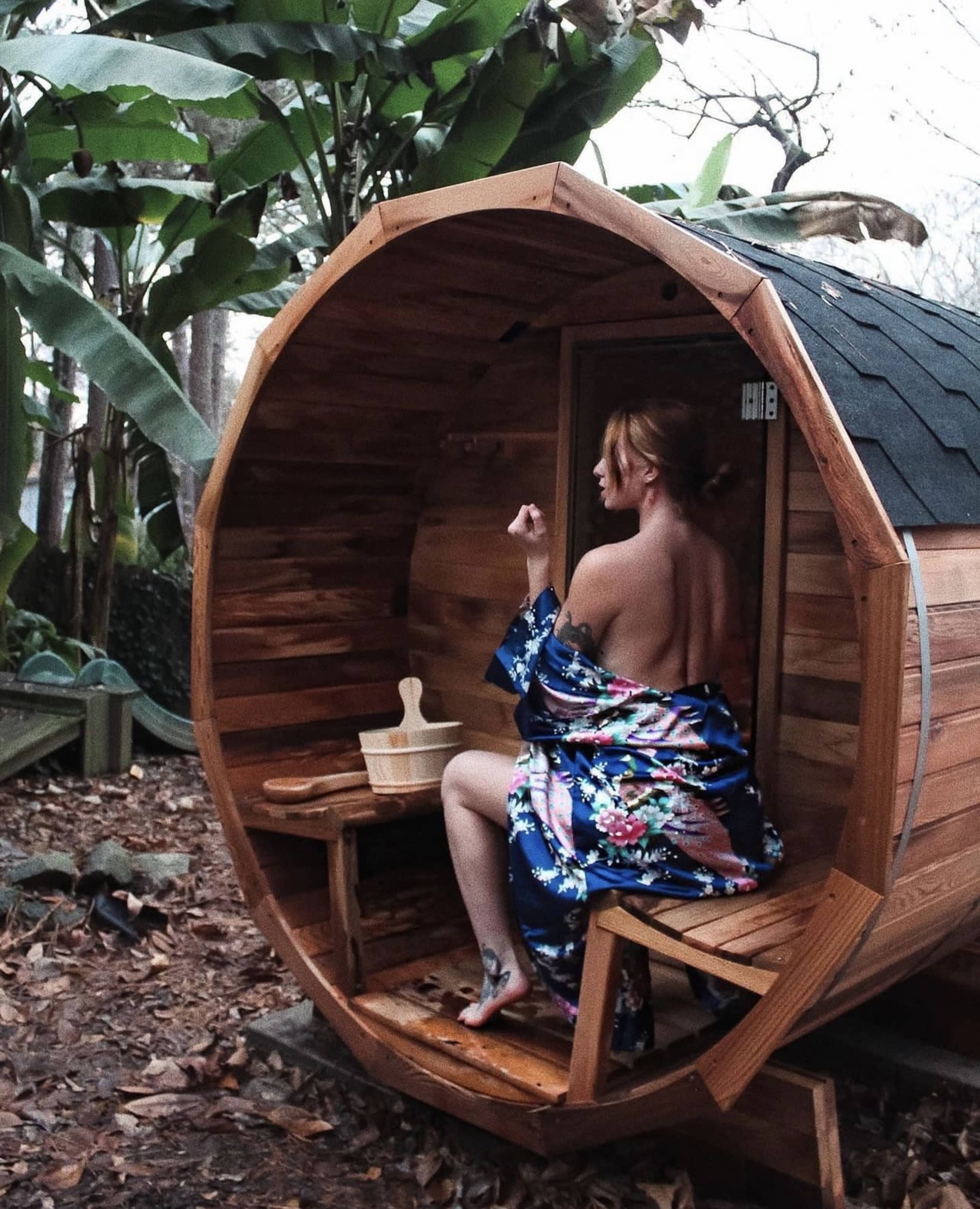 A person sits inside a wooden barrel sauna surrounded by tropical plants.