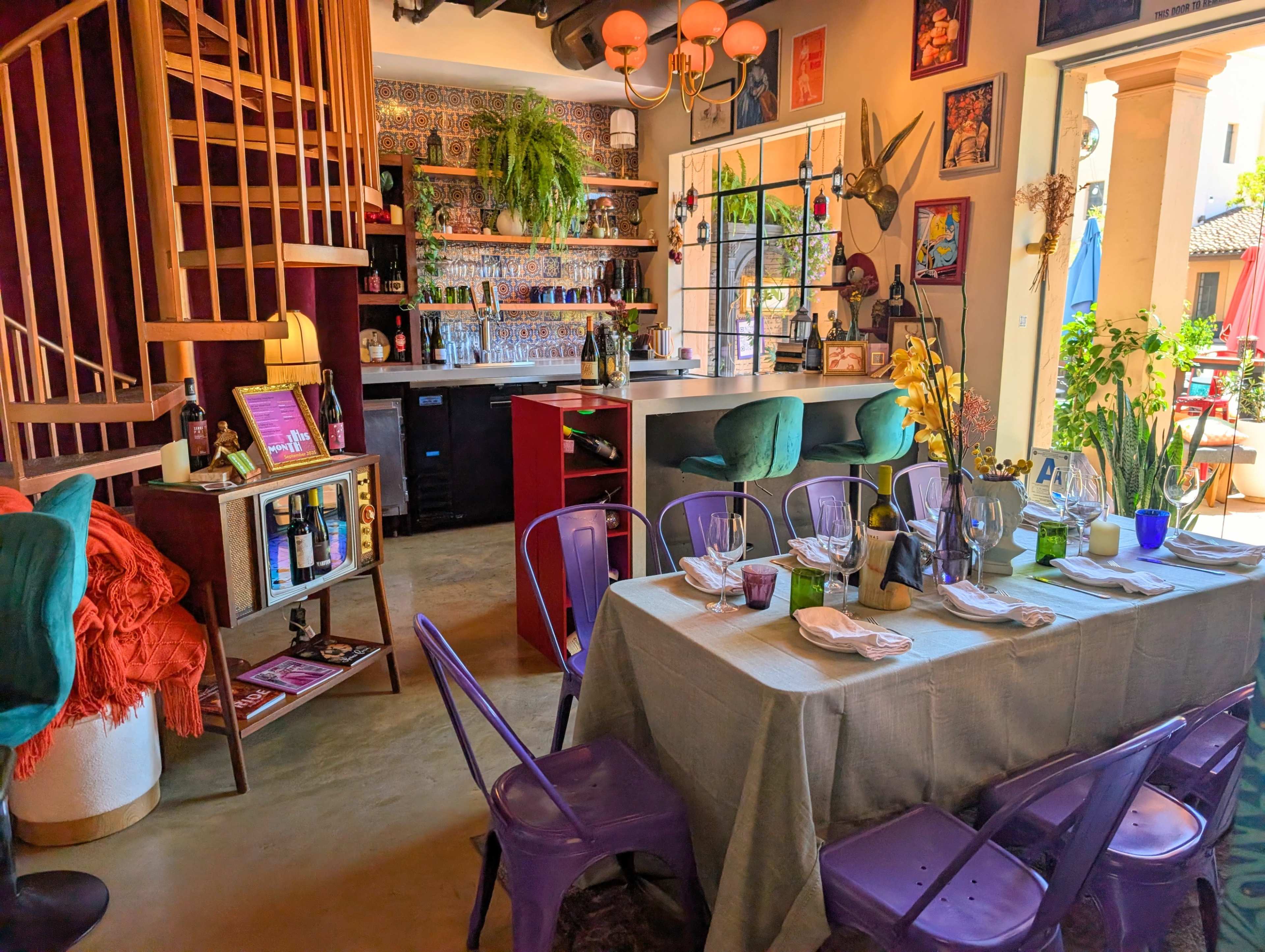 The image shows a colorful restaurant interior featuring a wooden spiral staircase, a bar area with various decorations, and a dining table set for a meal.