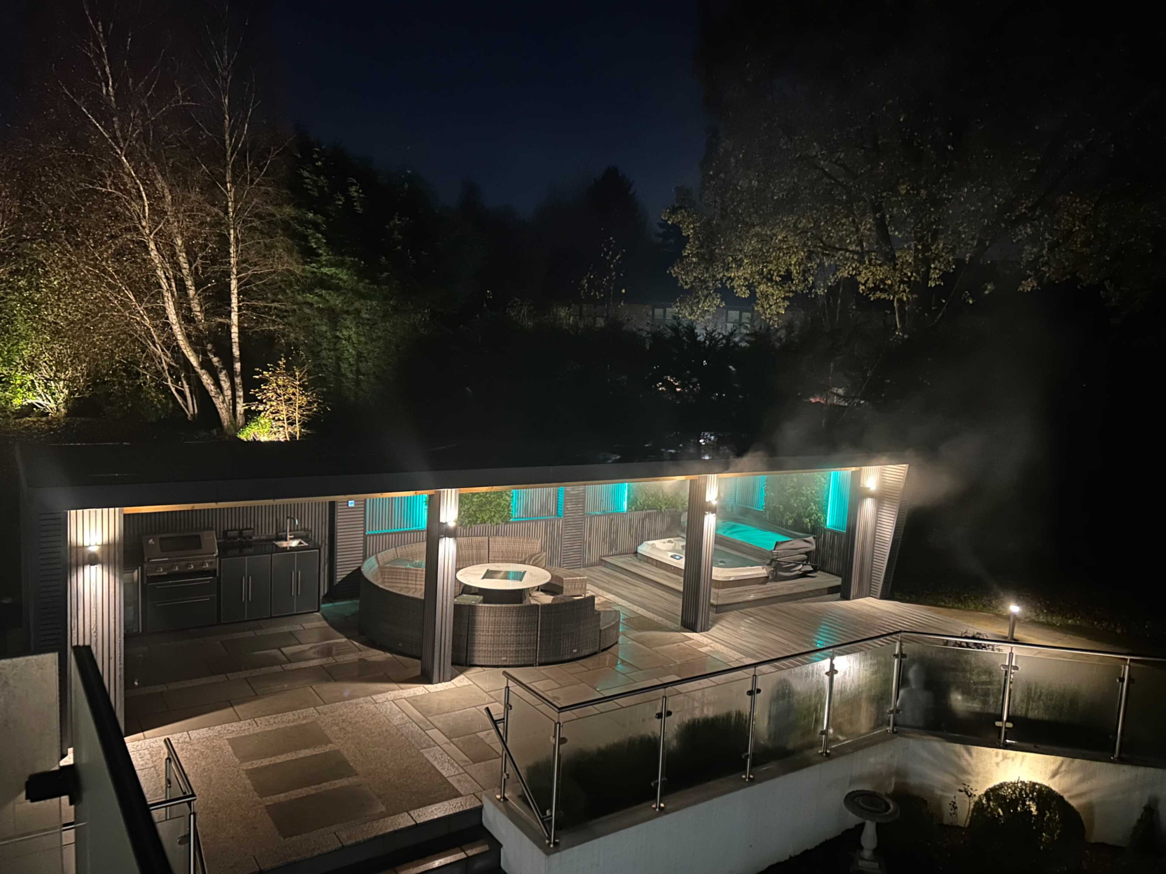 The image shows a modern outdoor area featuring a hot tub, a seating area, and ambient lighting amidst a nighttime setting.