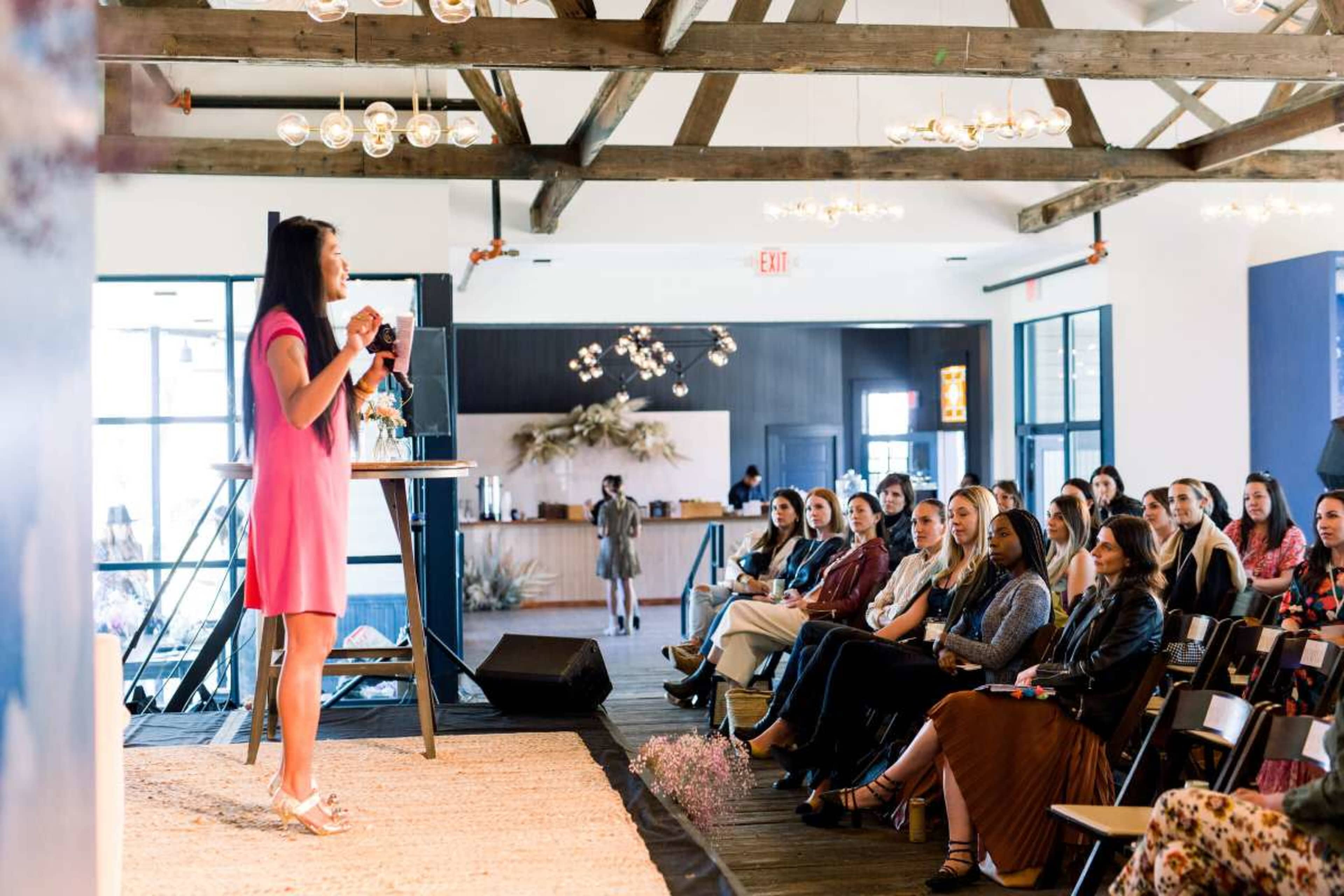 A speaker in a pink dress stands on stage addressing an audience seated in a well-lit room with large windows and wooden beams.