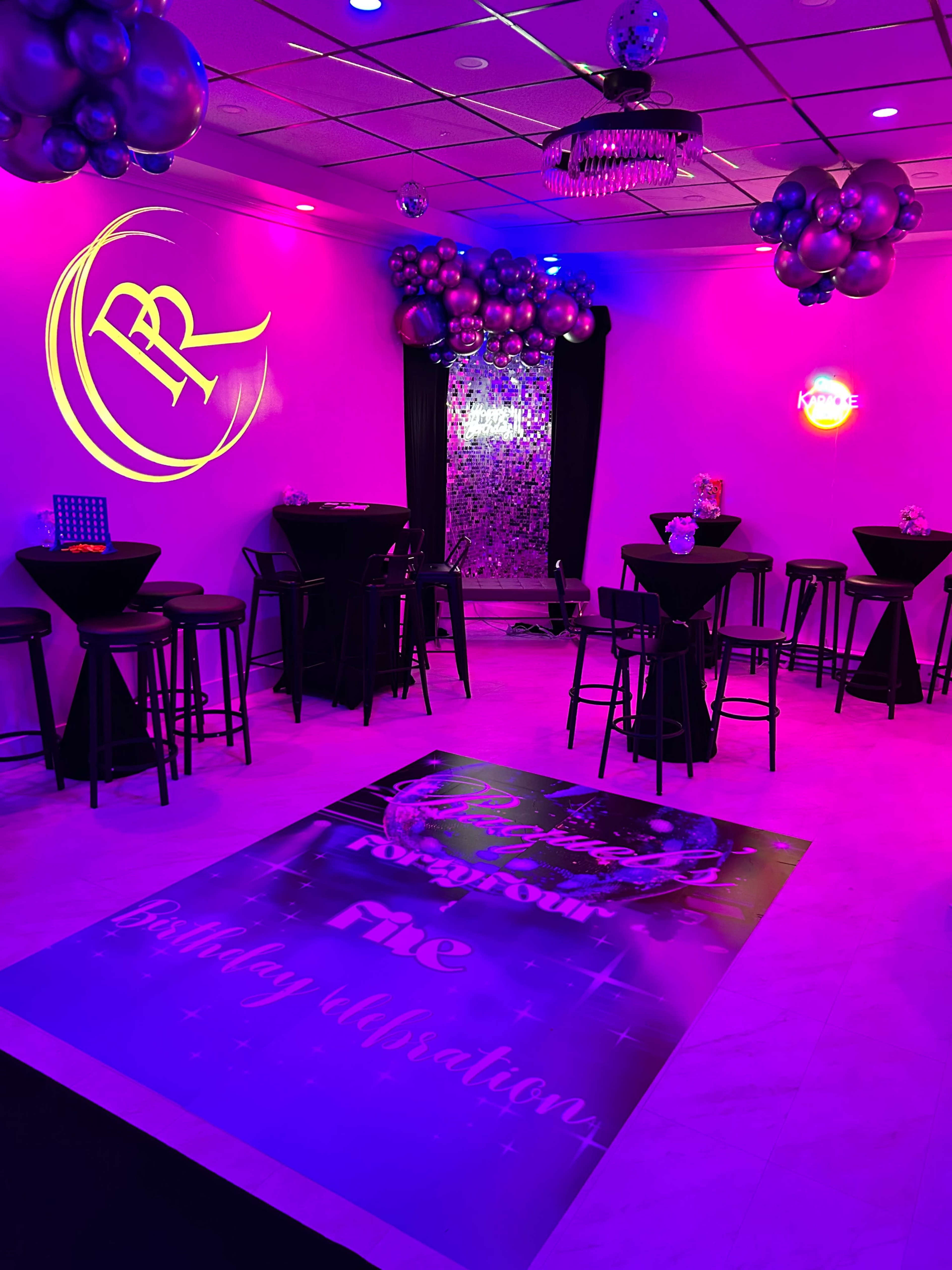 The image shows a vibrant party venue decorated with purple lighting, black tables, and a backdrop of balloons, featuring a large floor display that reads "Birthday Celebration."