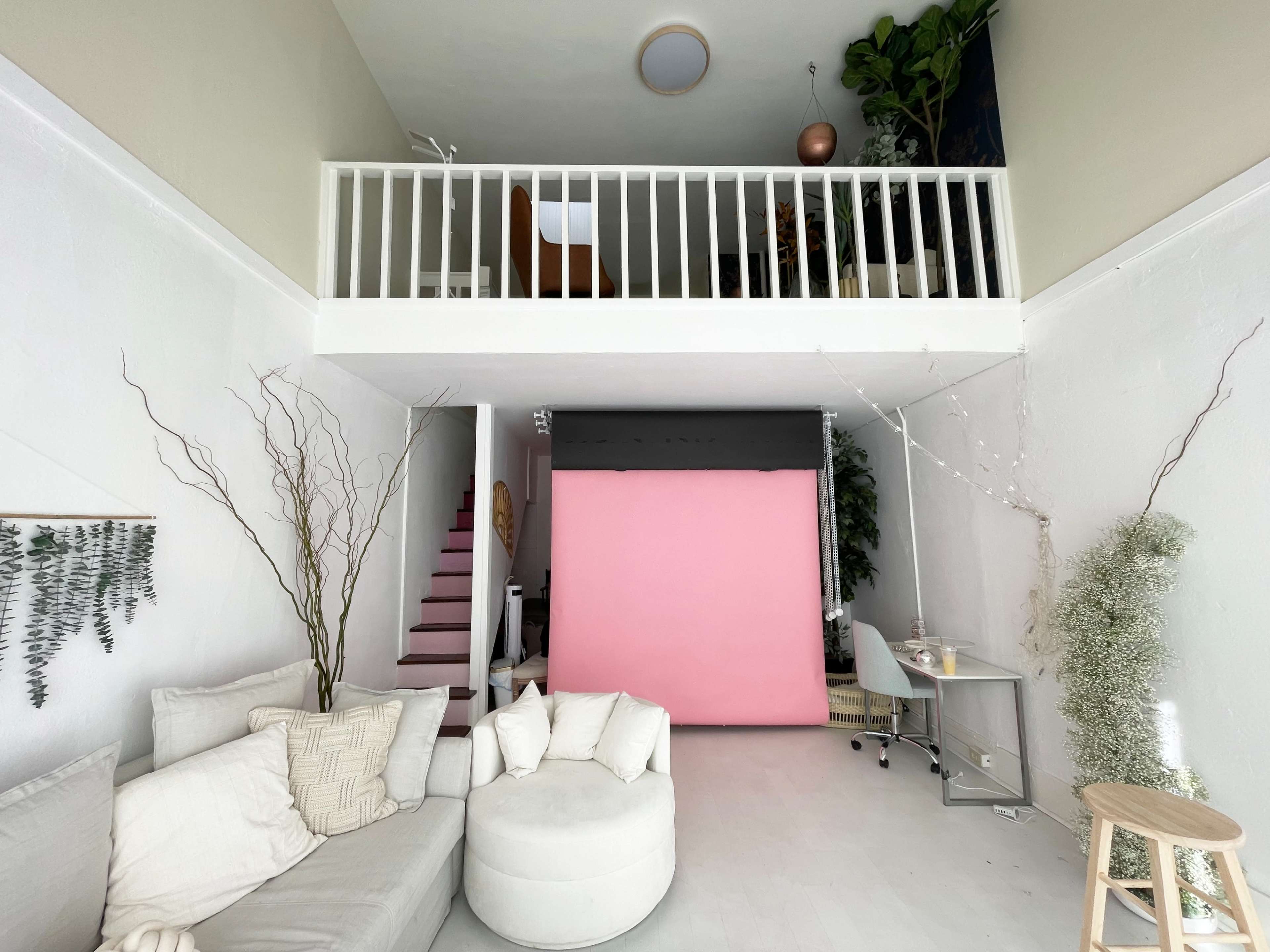 A spacious interior with a loft area, featuring a pink backdrop, a desk, a light-colored sofa, and decorative plants.