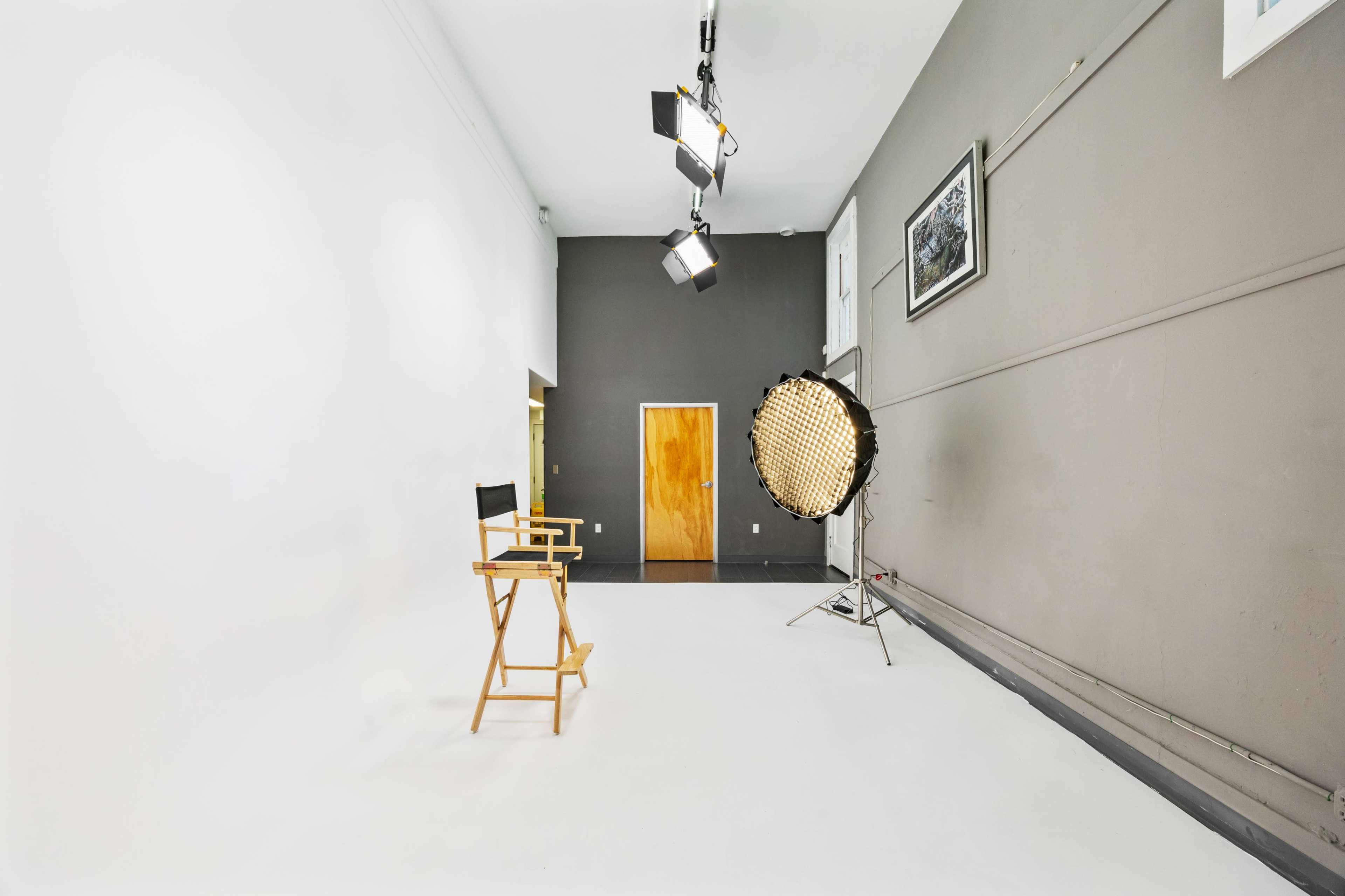 A photography studio features a wooden director's chair, a large light setup, and a white backdrop against a gray wall.