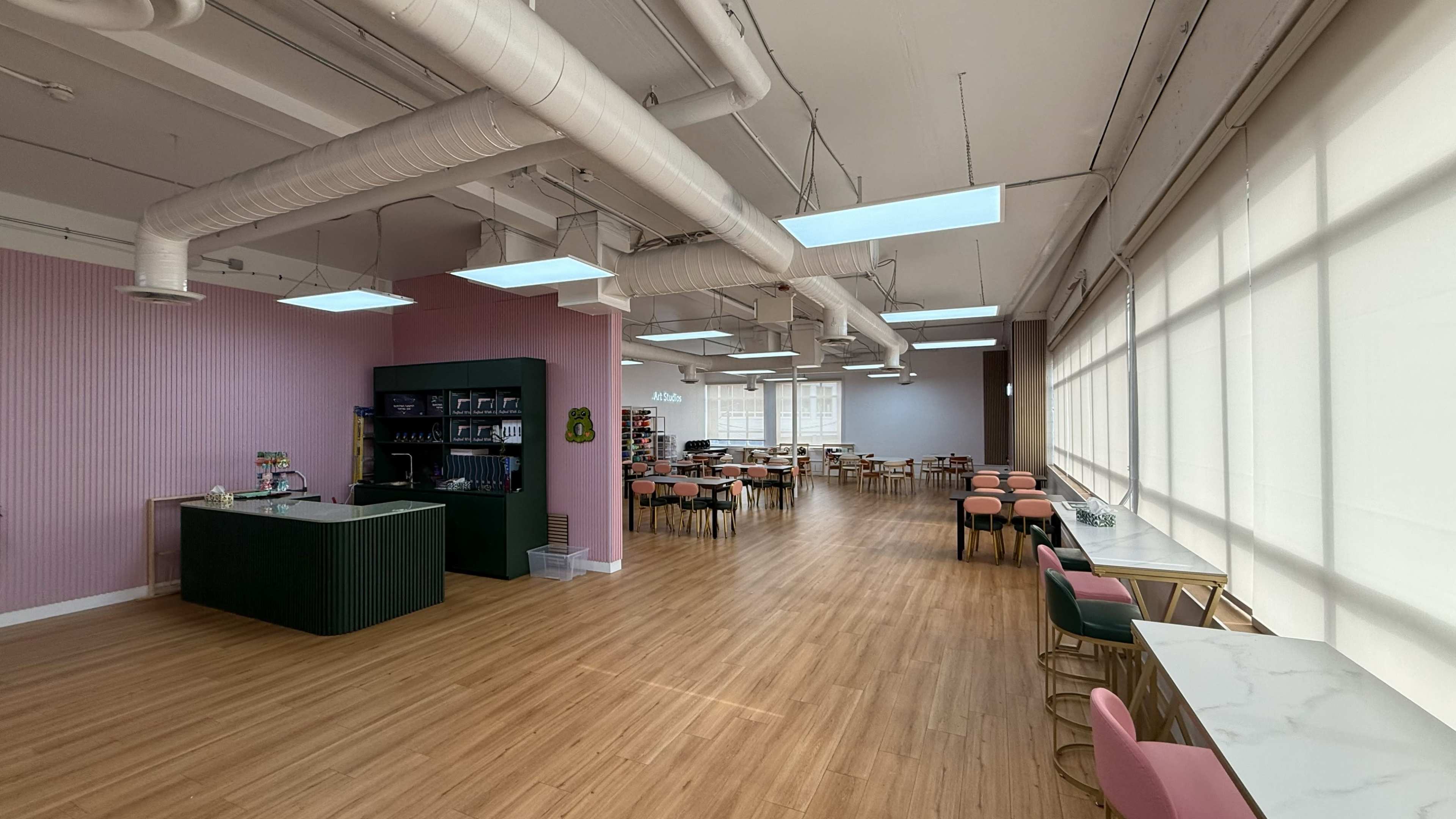 The image shows a spacious, modern café interior with pink walls, large windows, and various tables and chairs arranged throughout.