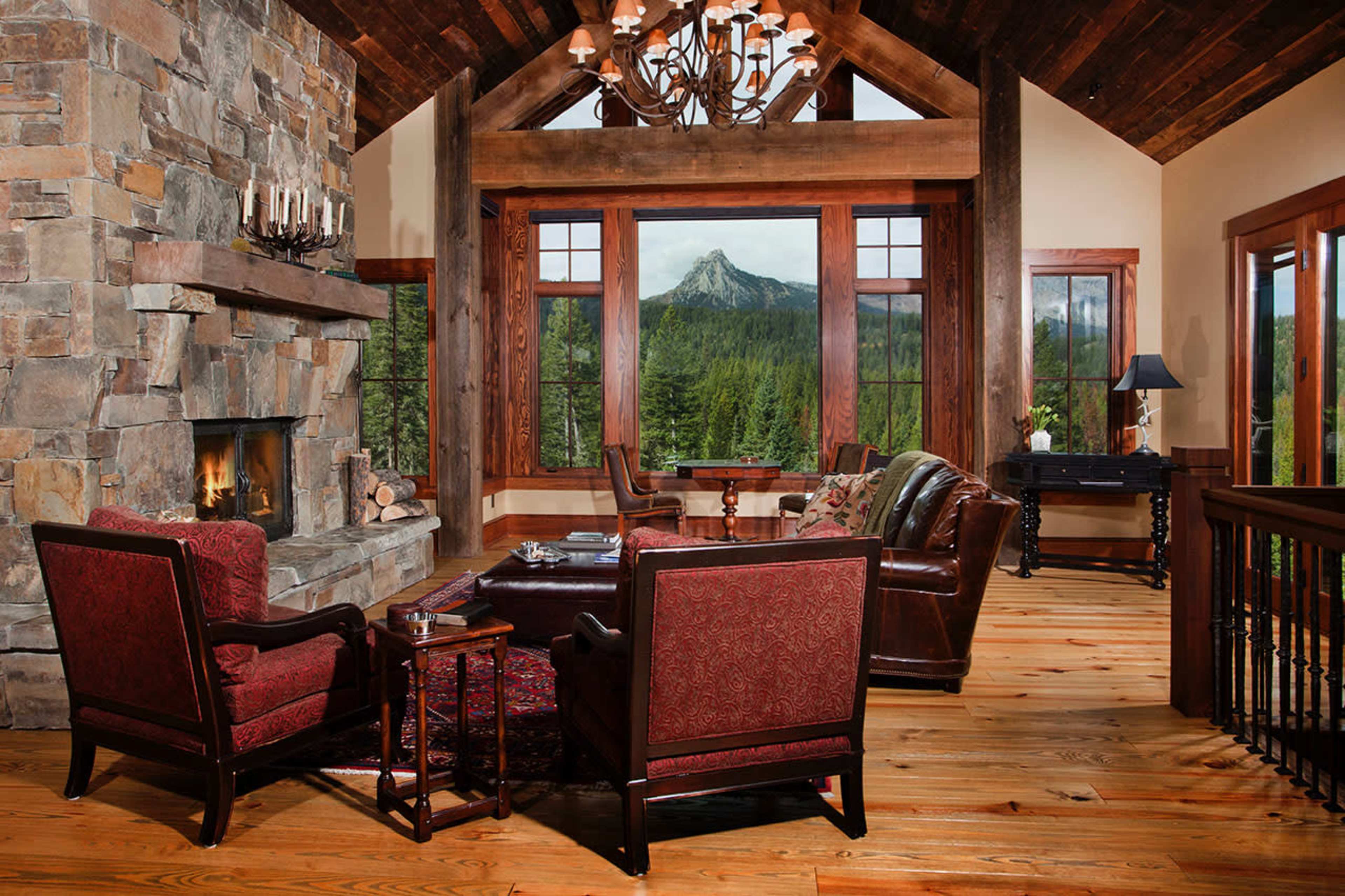 A spacious living room features a stone fireplace, large windows showcasing a mountain view, and leather seating arranged around a wooden coffee table.