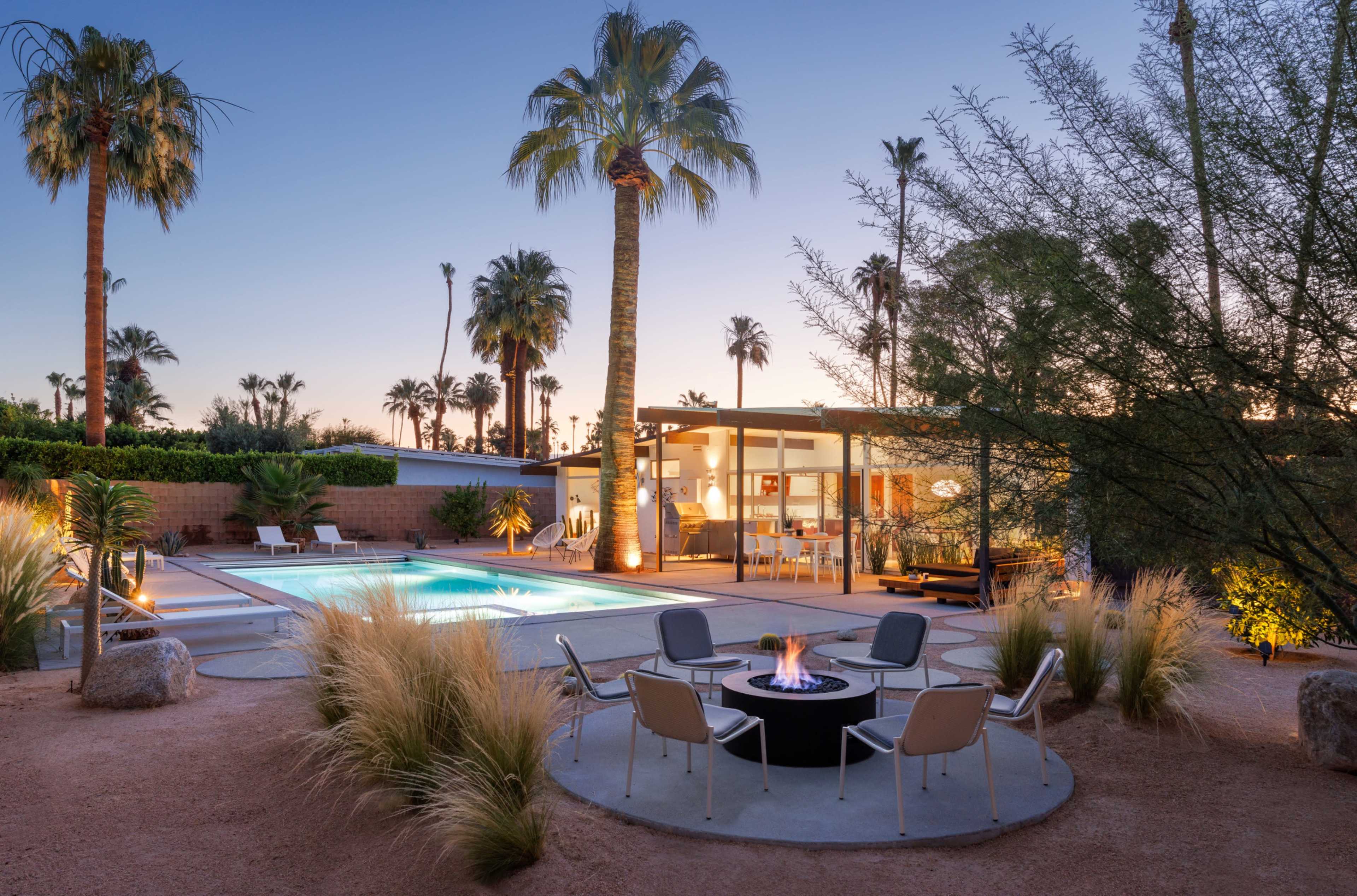 The image shows a modern backyard with a swimming pool, fire pit, and several palm trees at dusk.