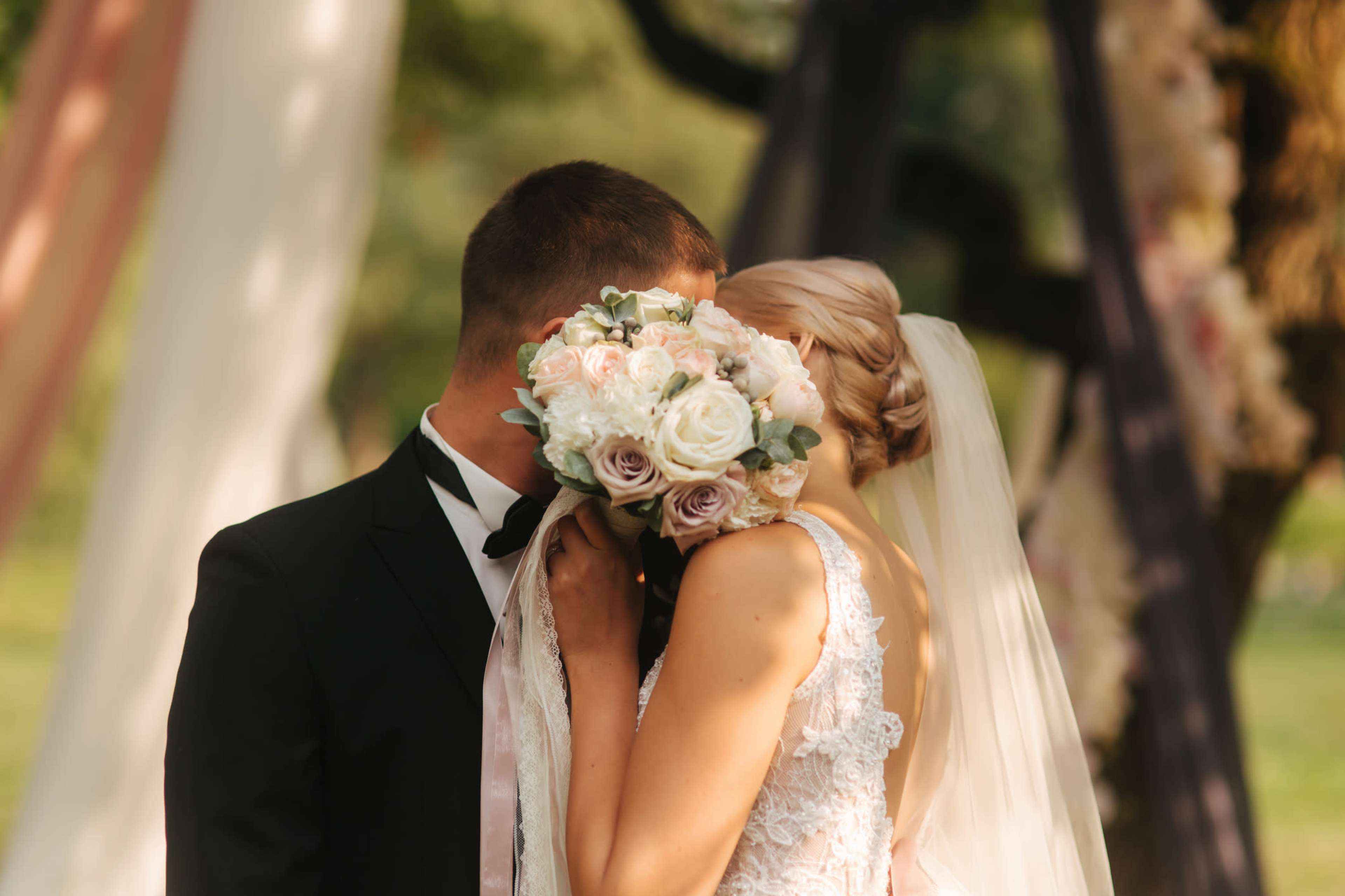 A bride and groom share a kiss while the bride holds a bouquet of roses, standing in front of a decorated outdoor ceremony backdrop.