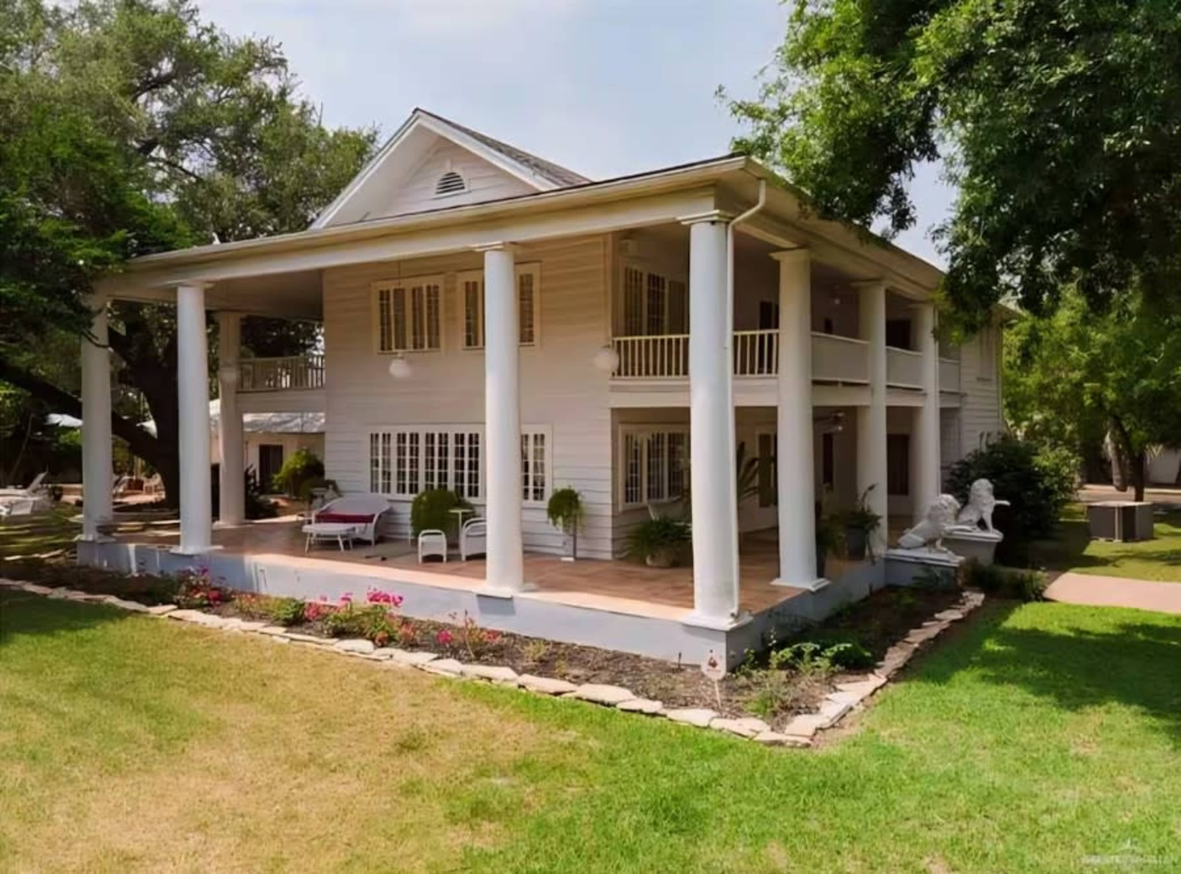 The image shows a two-story, white wooden house with large columns, surrounded by a green lawn and landscaped flowerbeds.