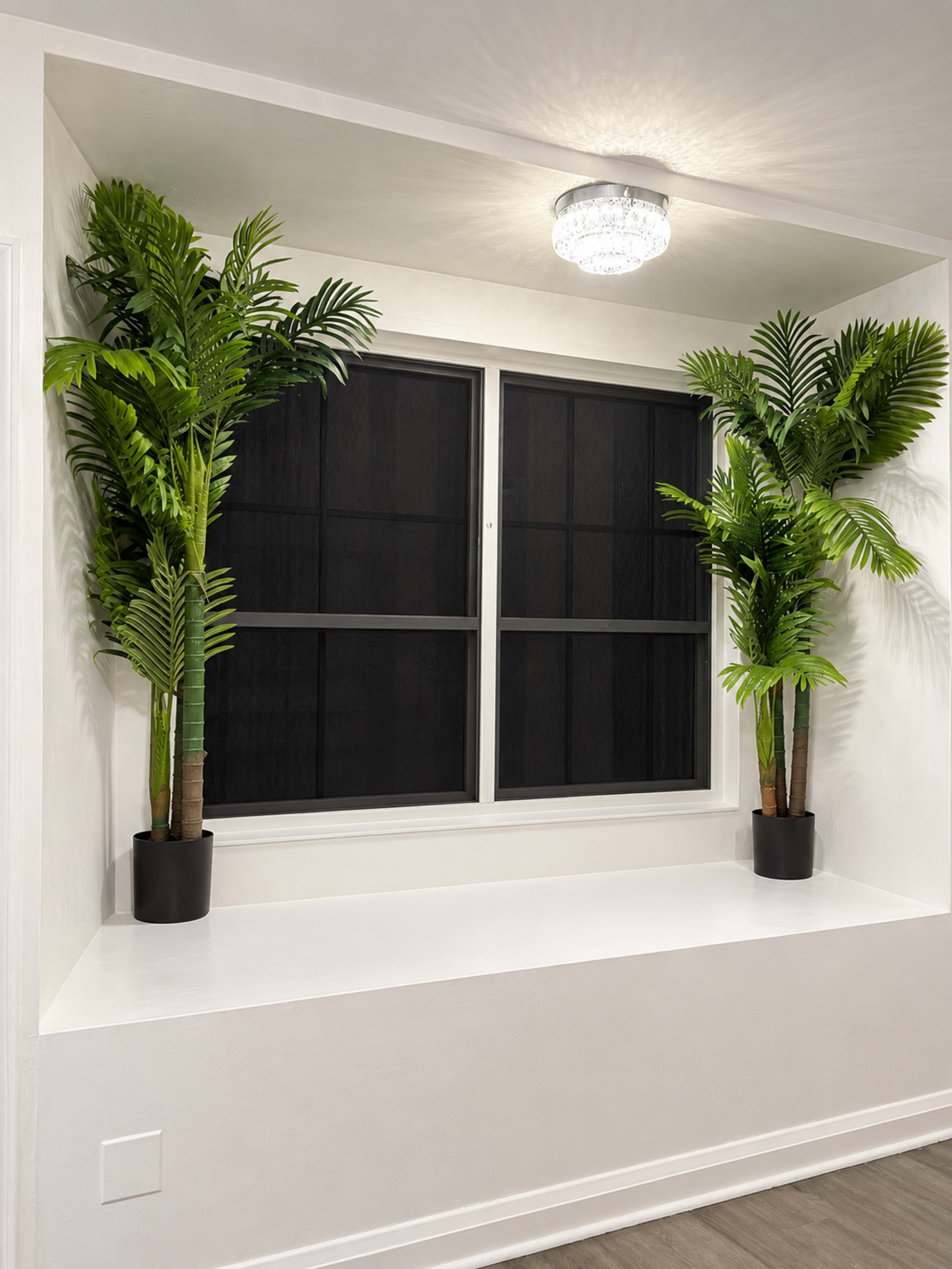 The image shows a window nook with two potted artificial palm plants flanking a rectangular window.