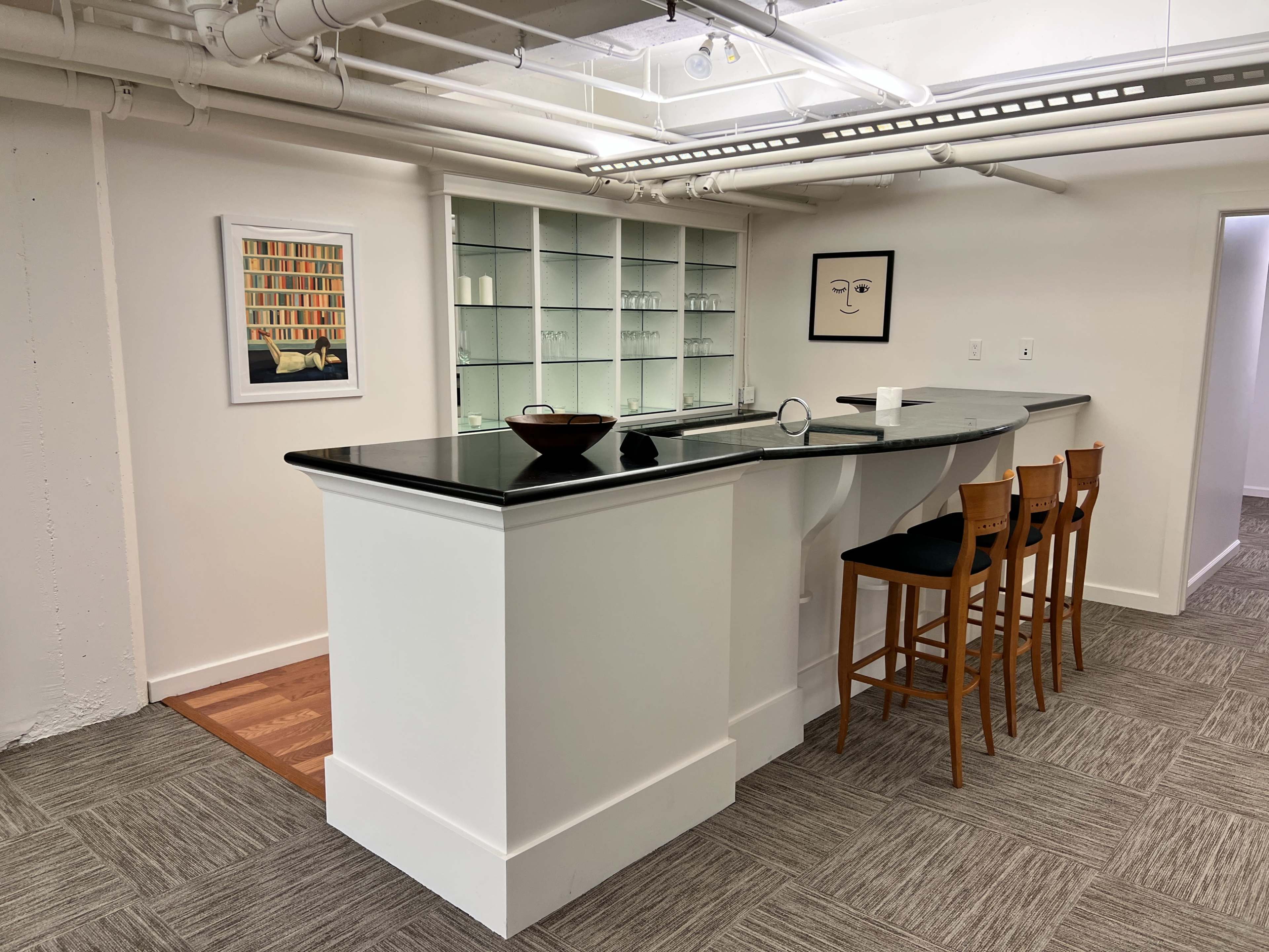 The image shows a modern, minimalist kitchen area with a black countertop, light-colored cabinetry, and wooden bar stools.