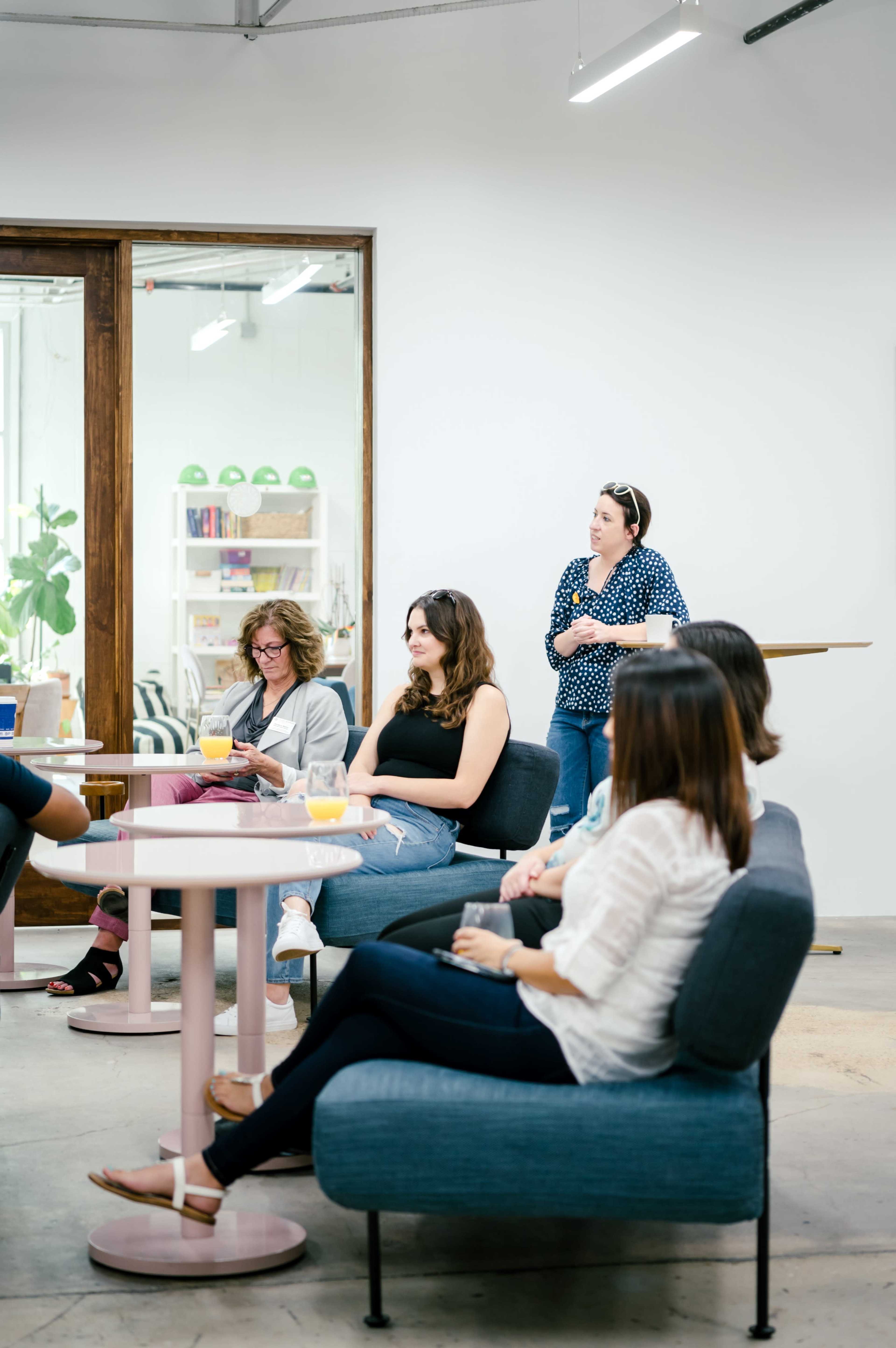 A group of women gathers in a modern, well-lit room with a casual seating arrangement, engaging in conversation.